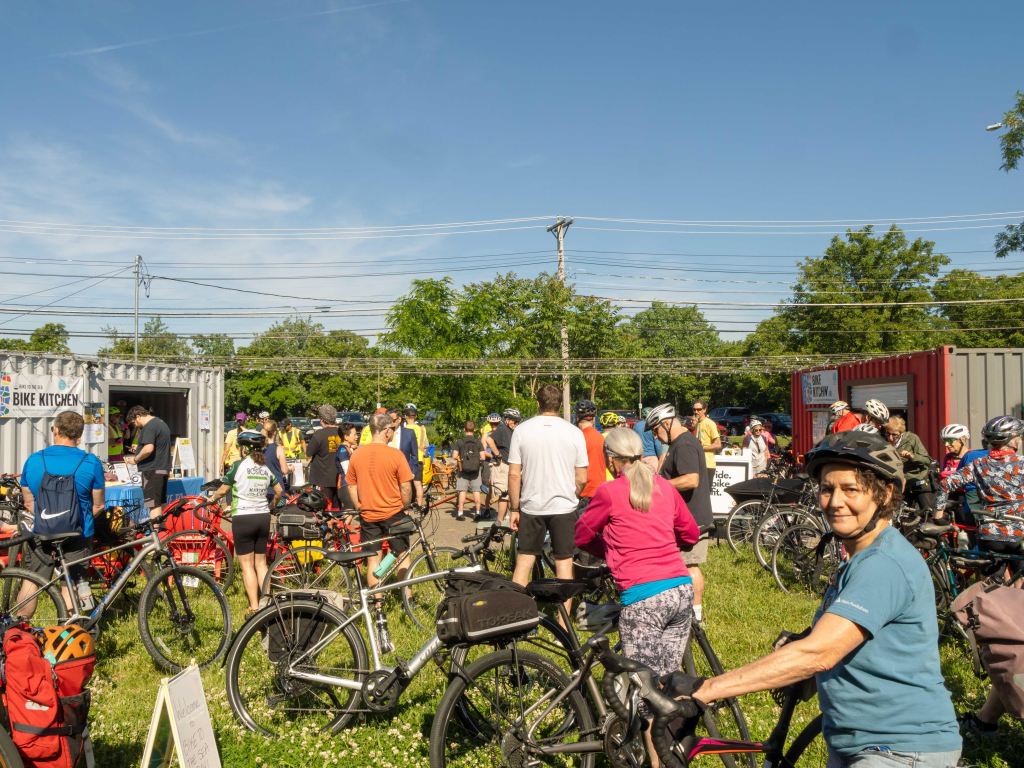 A large crowd of cyclists and their bikes in a field between a grey and a red shipping container.