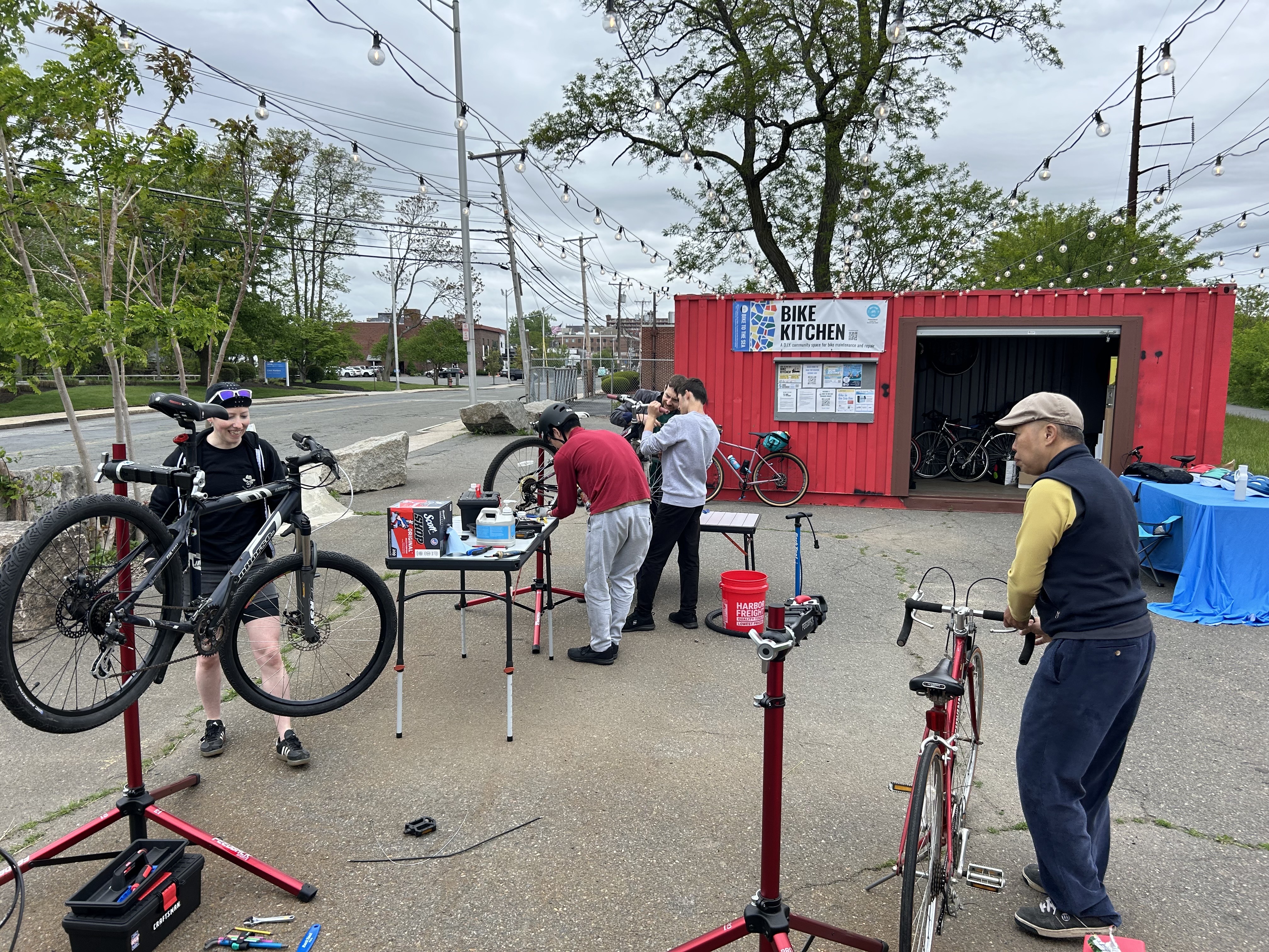 Several people working on bicycles mounted on red repair stands in a paved outdoor area in front of an open red shipping container.