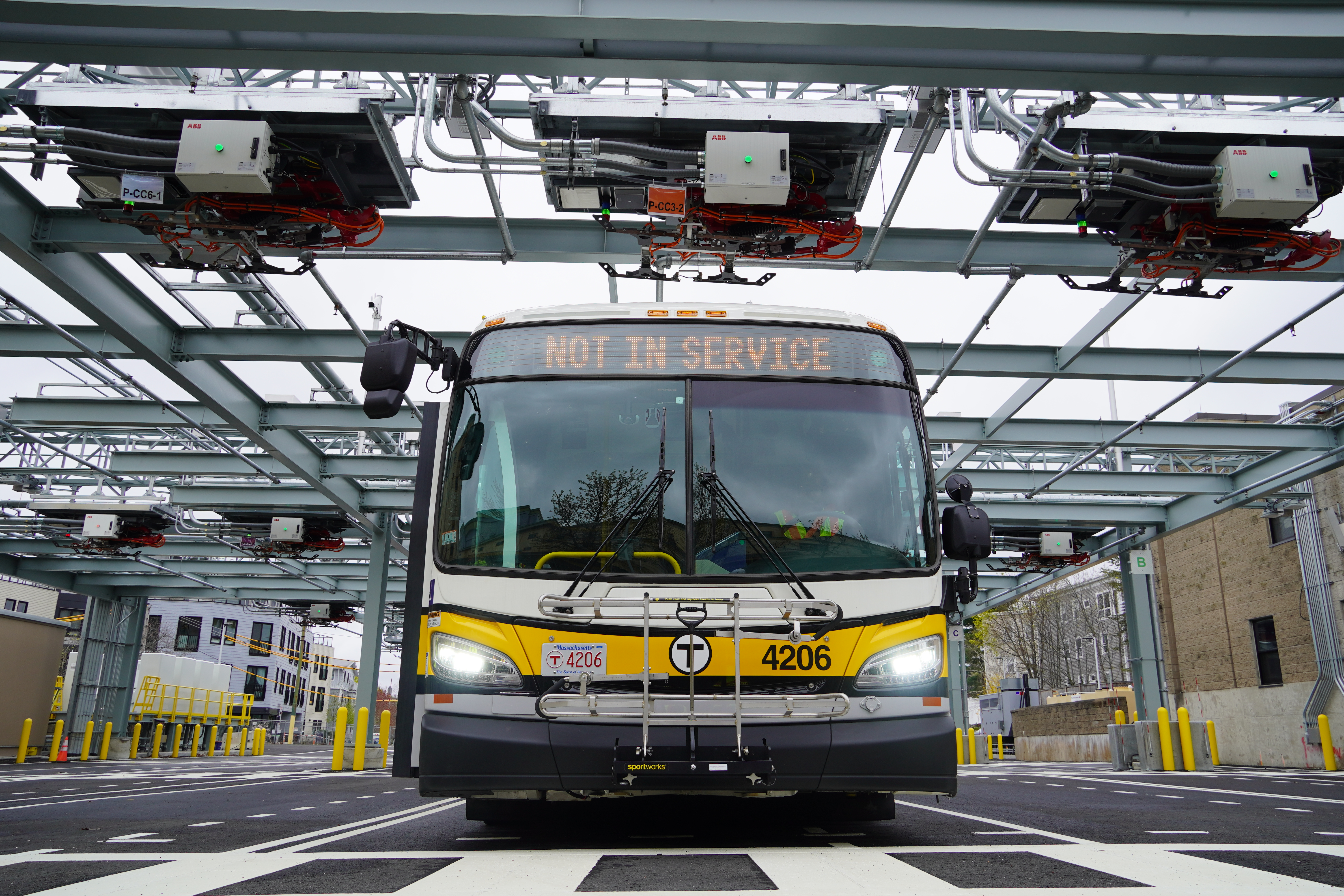 A ground-level view looking up at the front of MBTA bus 4206 with a "NOT IN SERVICE" sign. Above the bus is a large steel gantry of bus charging equipment.