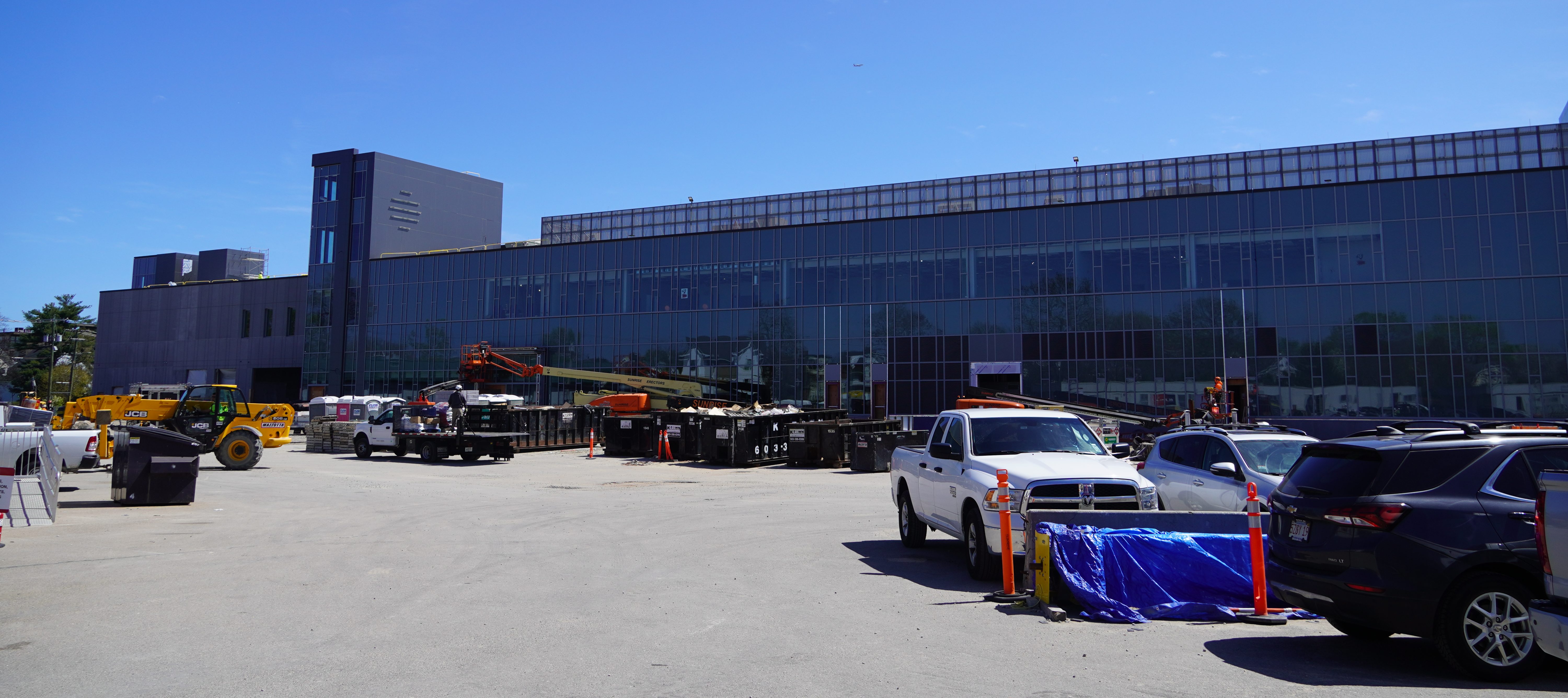 A large low-rise building clad in glass. In the foreground is a large parking lot with construction vehicles and dumpsters.
