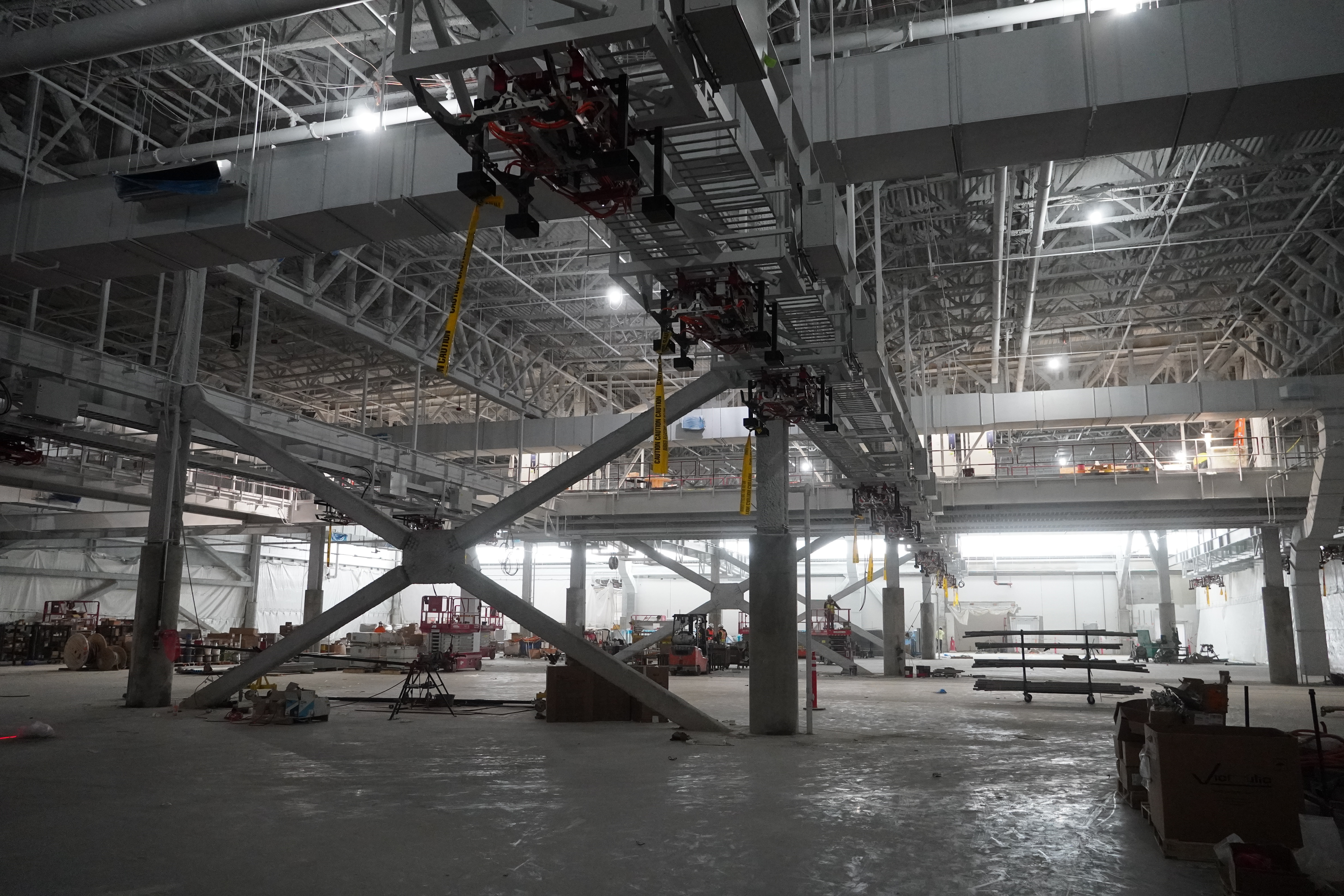 A huge warehouse-like building under construction. In the distance the far end of the hall is bathed in brighter sunlight from a skylight. The high ceilings are crowded with huge ducts. Below the ducts, a bus charging gantry hangs. The floor of the area is crowded with construction materials and lifts.
