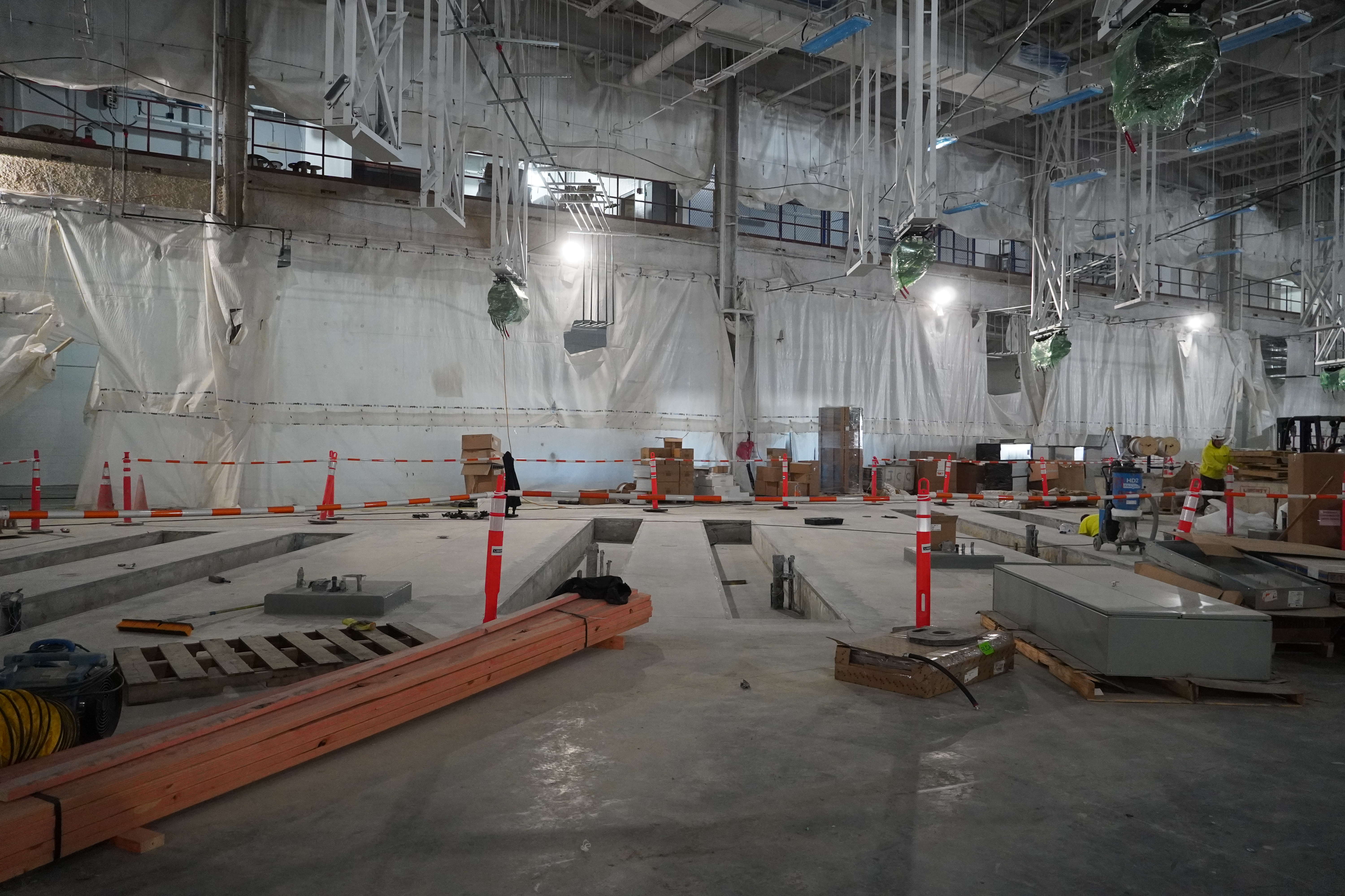 A huge warehouse-like building under construction. The high ceilings are crowded with huge ducts. The far wall is shrouded in plastic sheeting. In the center of the photo are several maintenance pits sunken into the concrete floor. Above, several charging gantries wrapped in green plastic descend from the ceiling.
