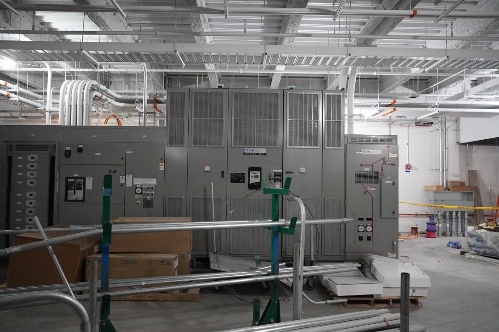 A huge gray cabinet of electrical equipment sits in a room scattered with construction materials and pipes.