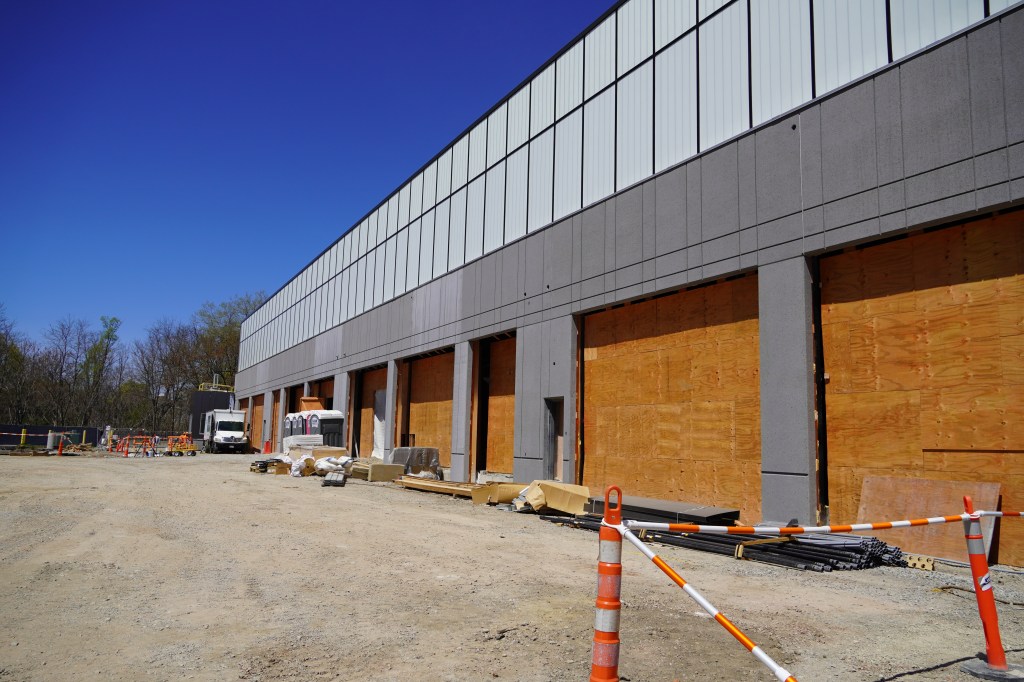 An exterior shot of a large gray building under construction with multiple garage bays covered in plywood and a gravel ground surface in front of the building.