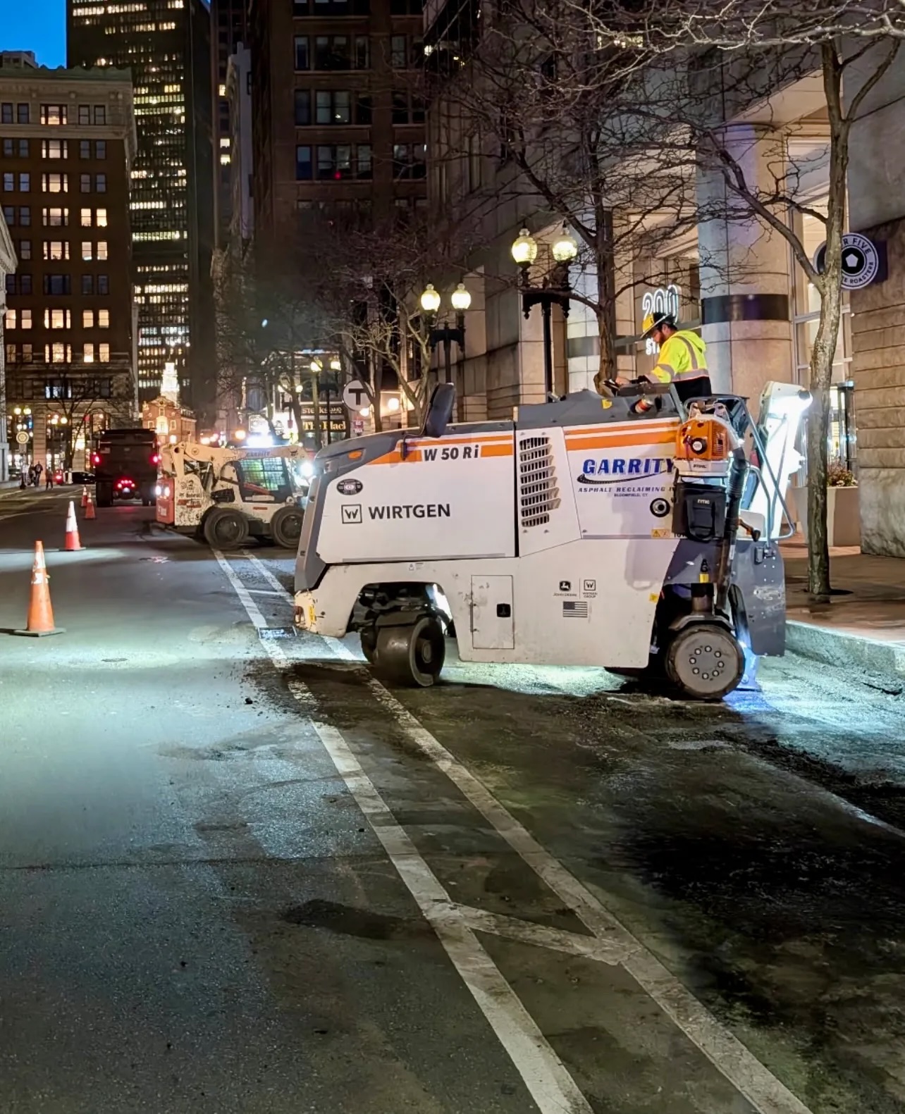 A construction vehicle navigates on top of a bike lane on a poorly-paved city street lined with tall buildings in the twilight.
