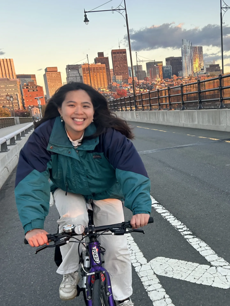 A woman with dark long hair in a teal and navy blue jacket and white pants smiling while riding a purple bicycle on a bridge with the Boston city skyline in the background.