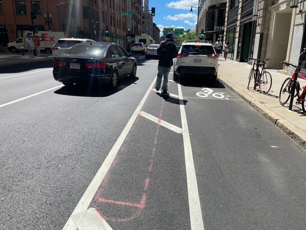 A photo of a bike lane blocked by a parked car on a newly paved city street. Spray-painted orange outlines between the bike lane and the car lane indicate the location of planned new concrete curbs. A skateboarder is merging into the car lane to get around the illegally parked vehicle.