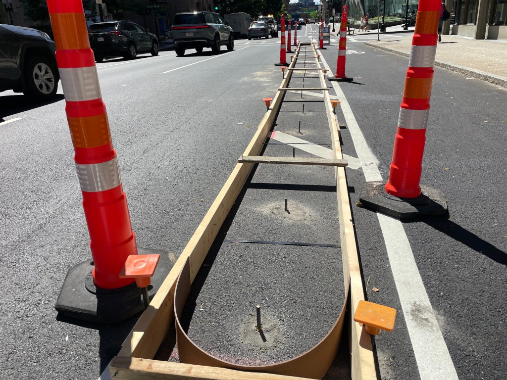 A photo of wooden concrete forms on top of a newly paved city street surrounded by orange construction cones.