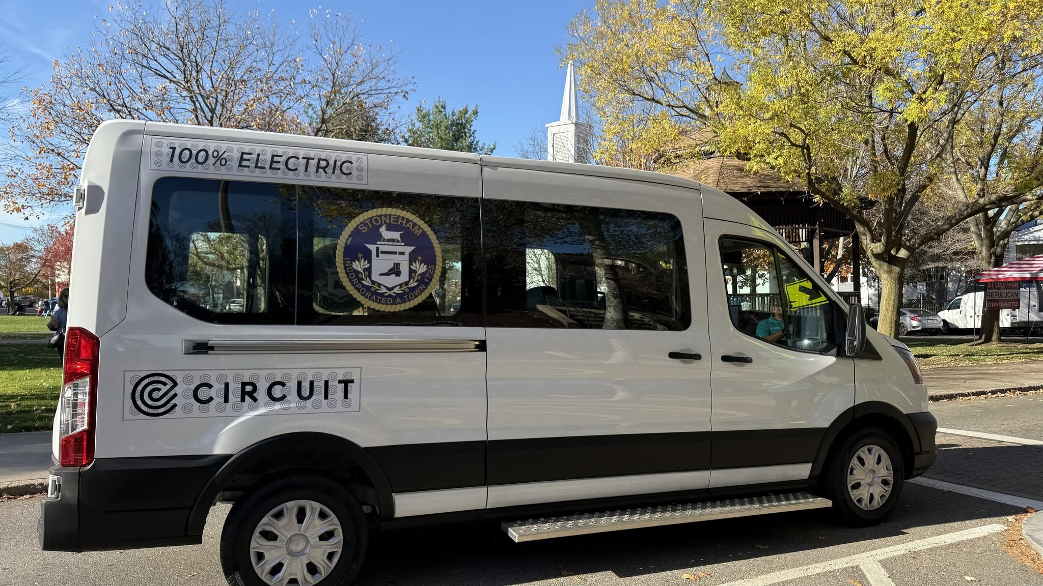 A large white passenger van is parked in front of a park gazebo with a white church steeple in the background. The side of the van bears the town seal of Stoneham, MA, and a banner above the window says "100% electric."