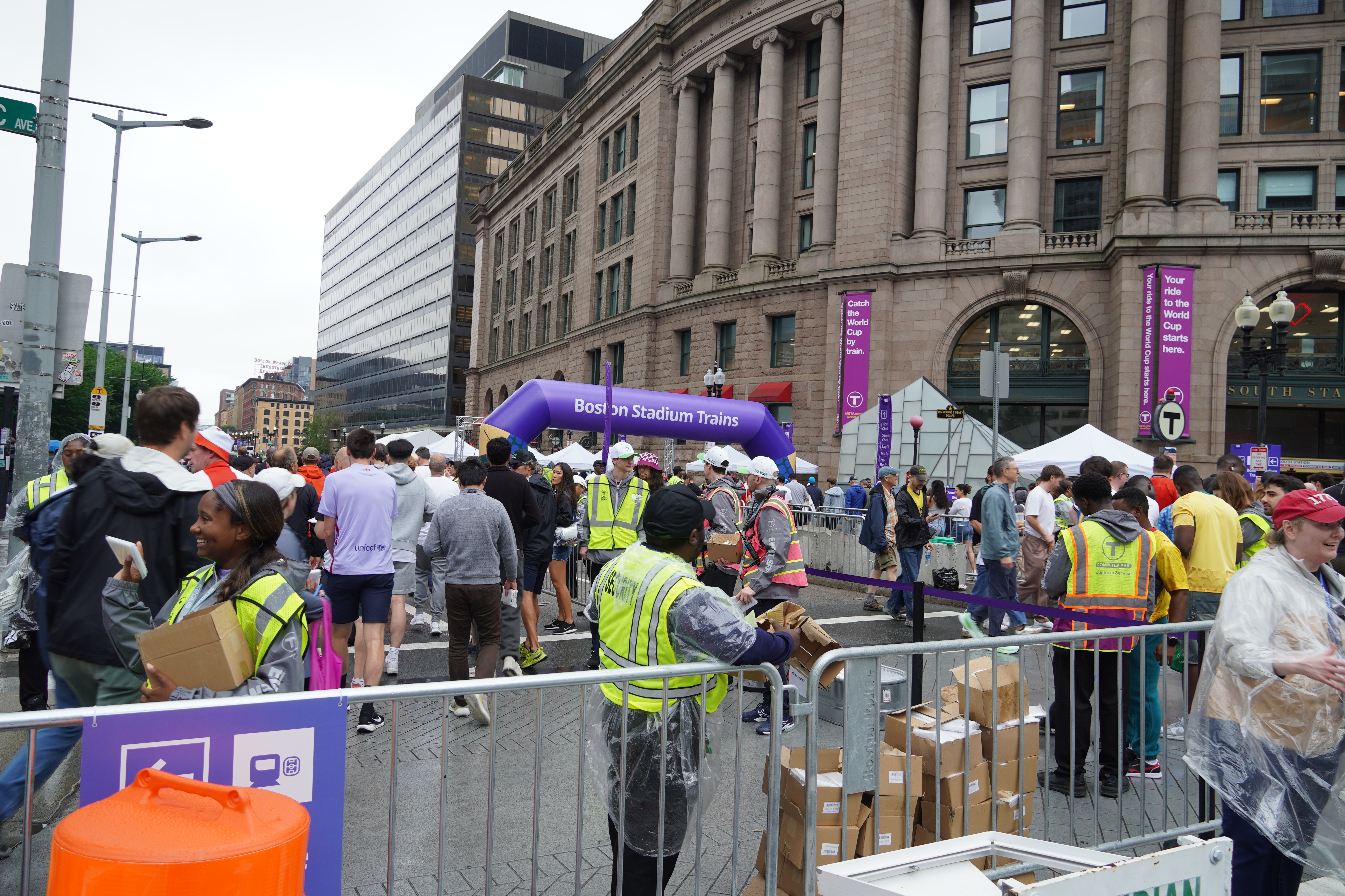 A crowd of people, several of whom are wearing flourescent yellow vests, mills around a closed-to-traffic street in front of Boston South Station. A purple inflated arch in the background reads "Boston Stadium Trains"