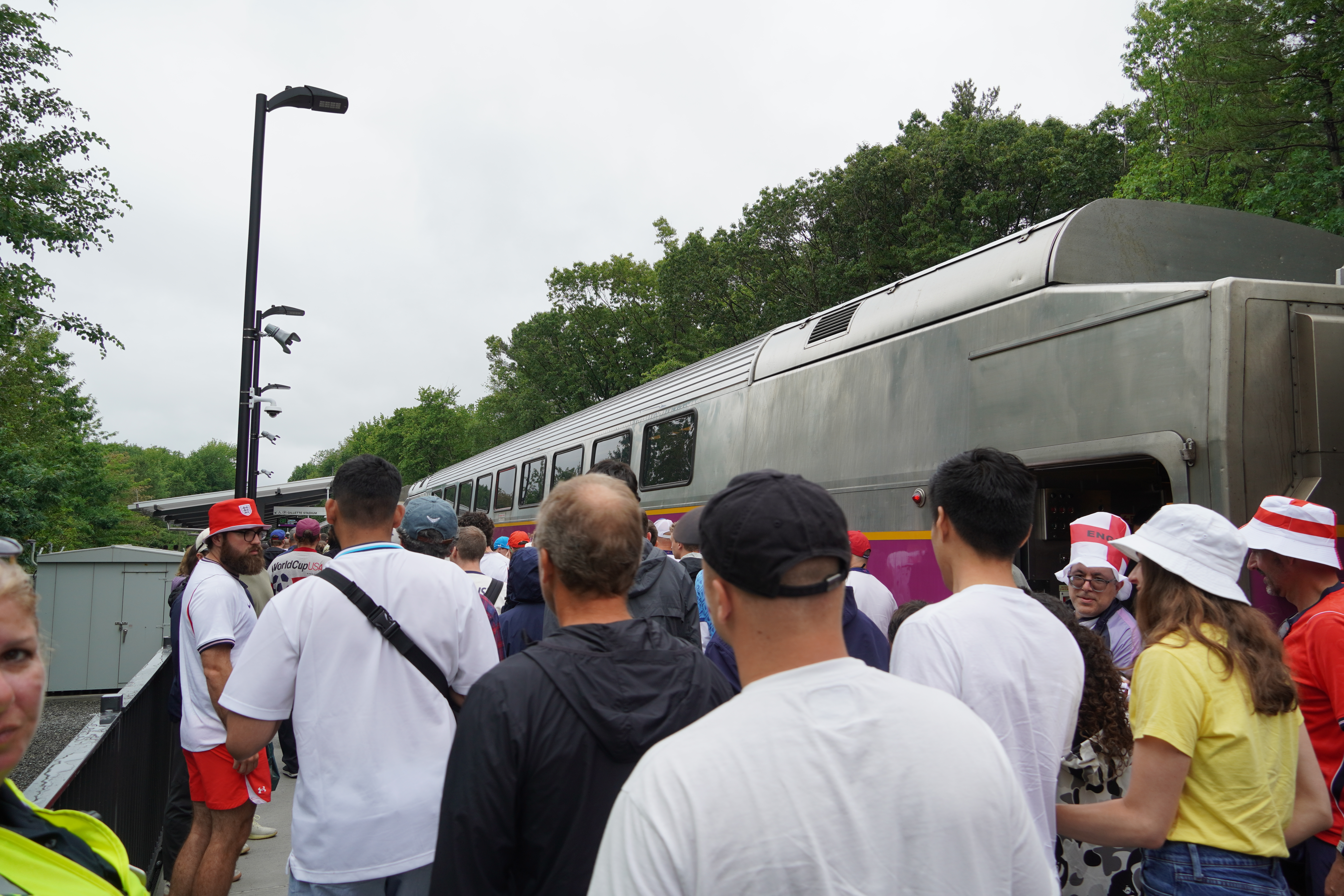 A crowd of soccer fans lines up next to an MBTA train on a platform surrounded by trees.