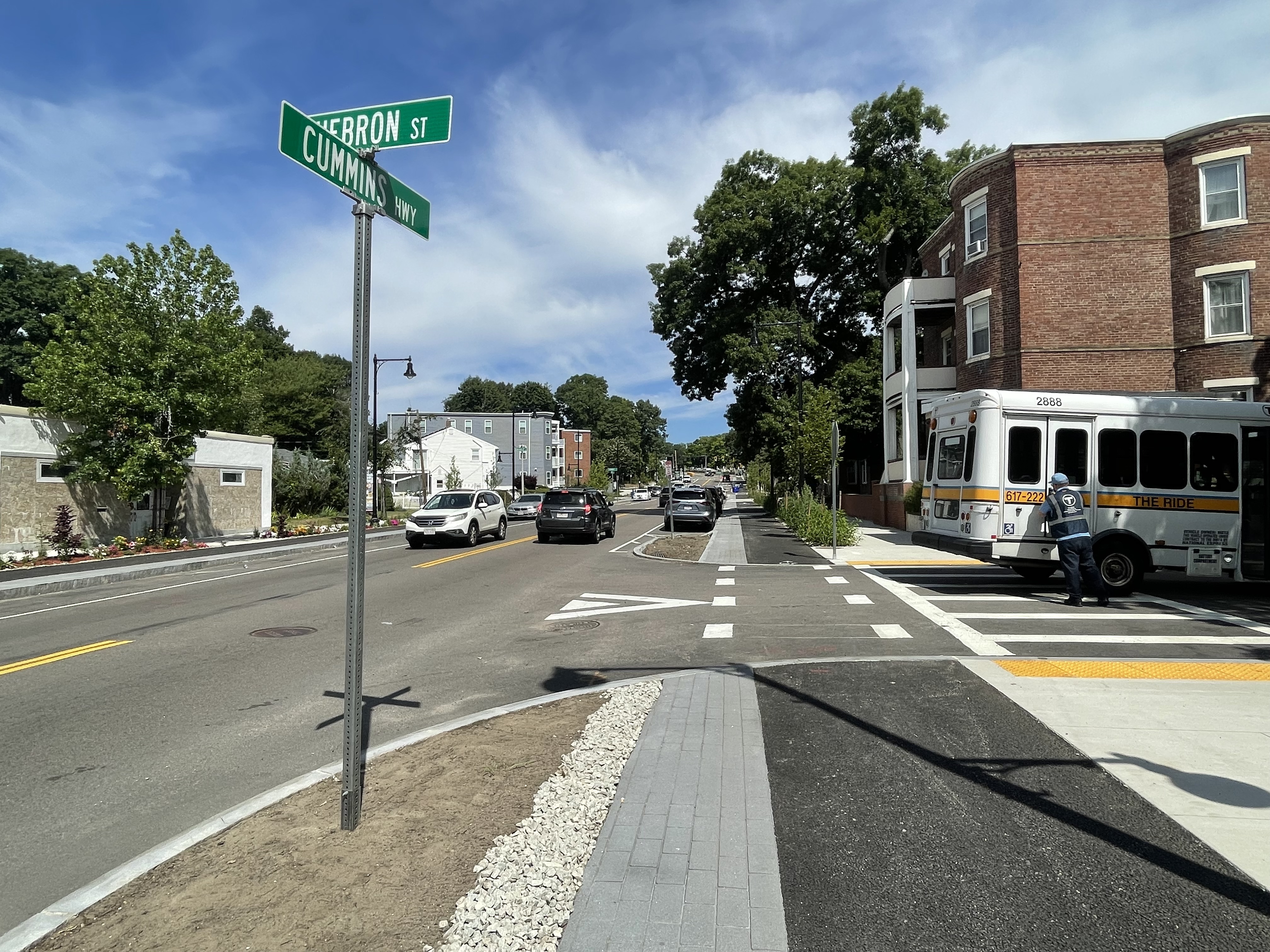 A view of a protected bike lane alongside a newly rebuilt city street with an MBTA "The Ride" van parked in a crosswalk on the adjacent side street. A street sign in the left foreground reads "Cummins Hwy" and "Hebron St" for the cross street.