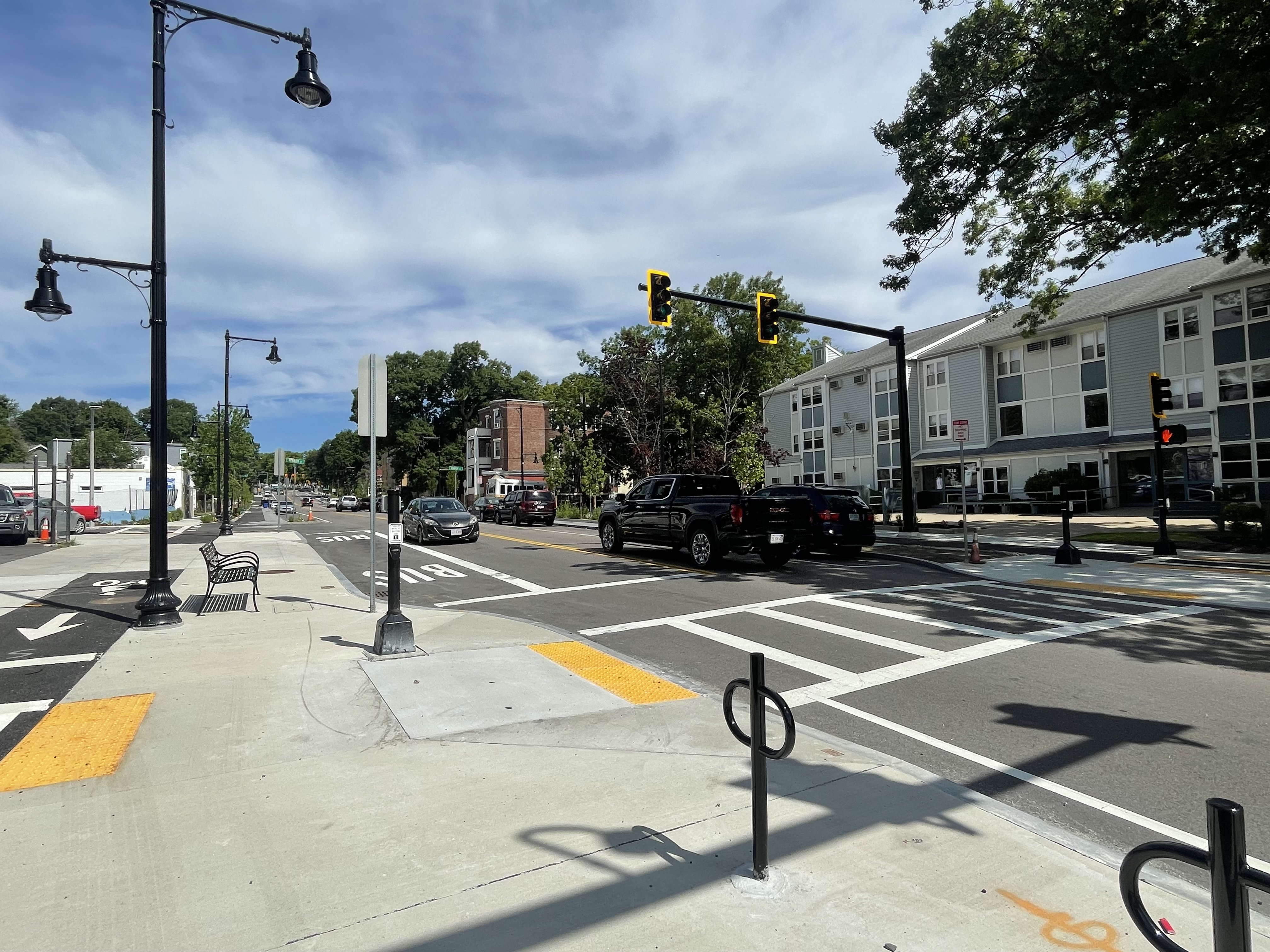 A view of a newly-built street with bike racks and a wide sidewalk in the foreground, and a traffic signal above a crosswalk in the center of the image. To the left is a bus stop and a bike lane behind a streetlamp and. bench. The street is lined with 3-story small apartment buildings and trees.