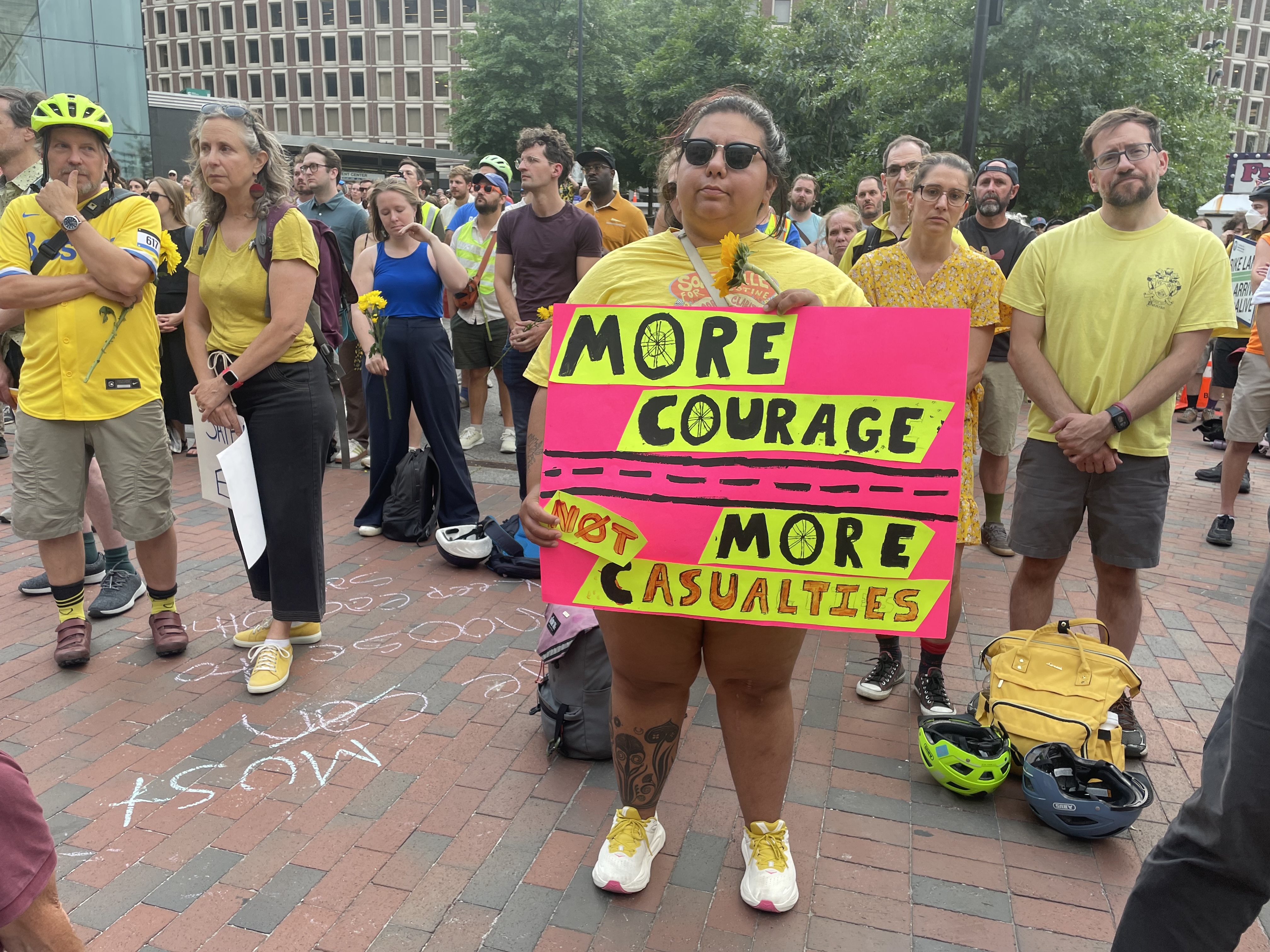 Somber-faced mourners stand in a wide brick plaza. The person in the front of the photo is wearing sunglasses and holding a large poster in front of them that reads "More courage, not more casualities"