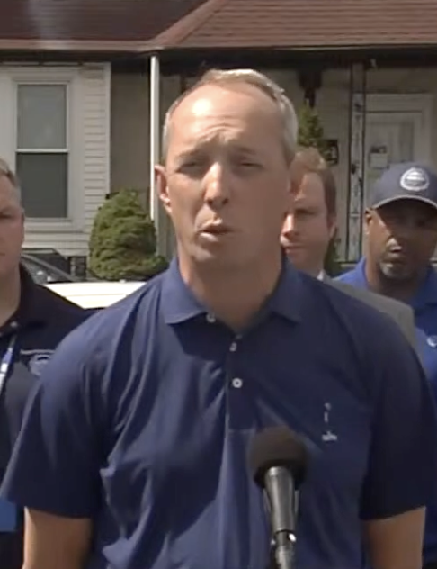 A middle-aged man in a blue polo shirt stands behind a microphone