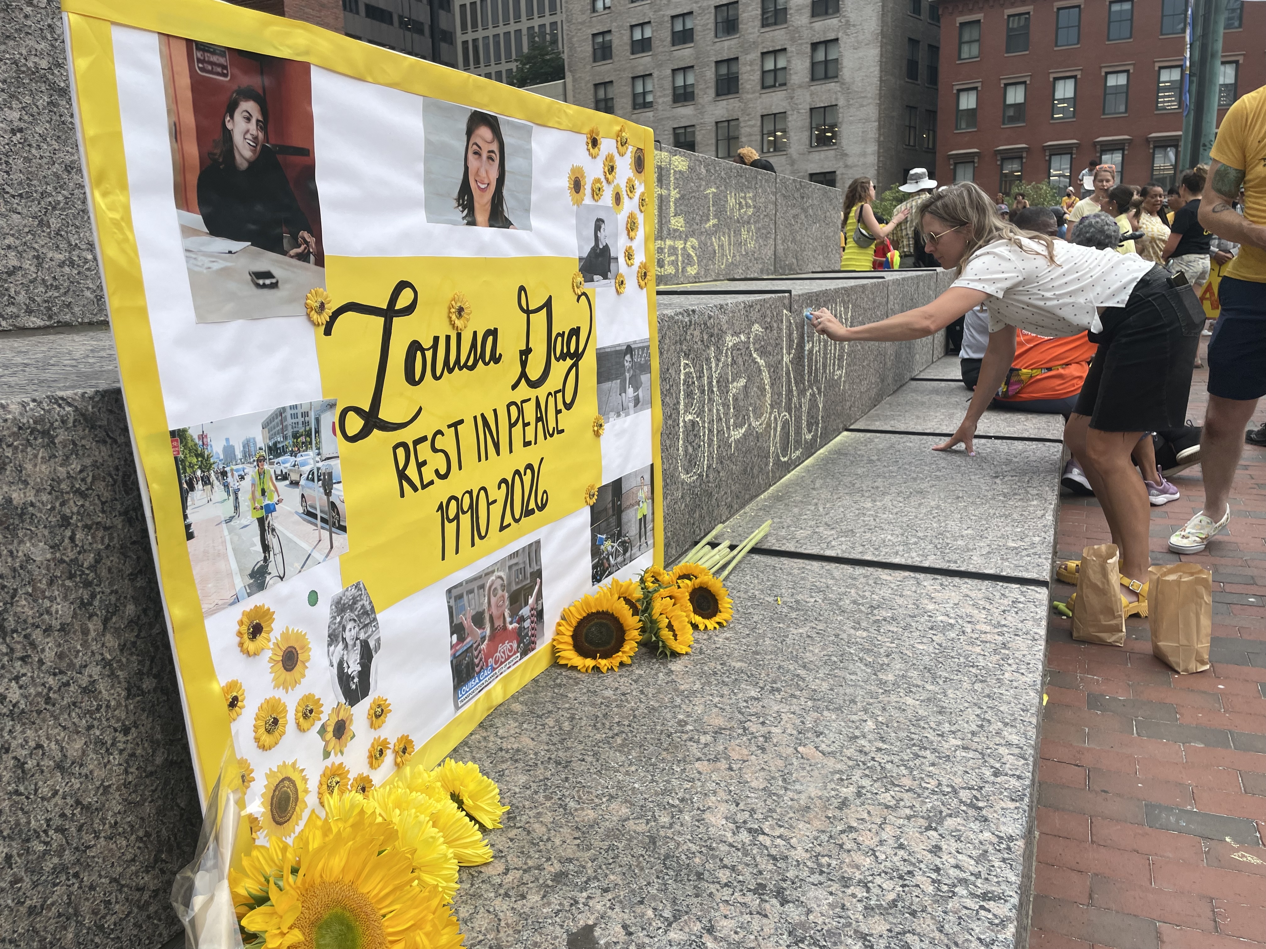 A poster with photos of a young woman sits on a granite ledge in front of some cut sunflowers. Text on the poster reads "Louisa Gag 1990-2026"