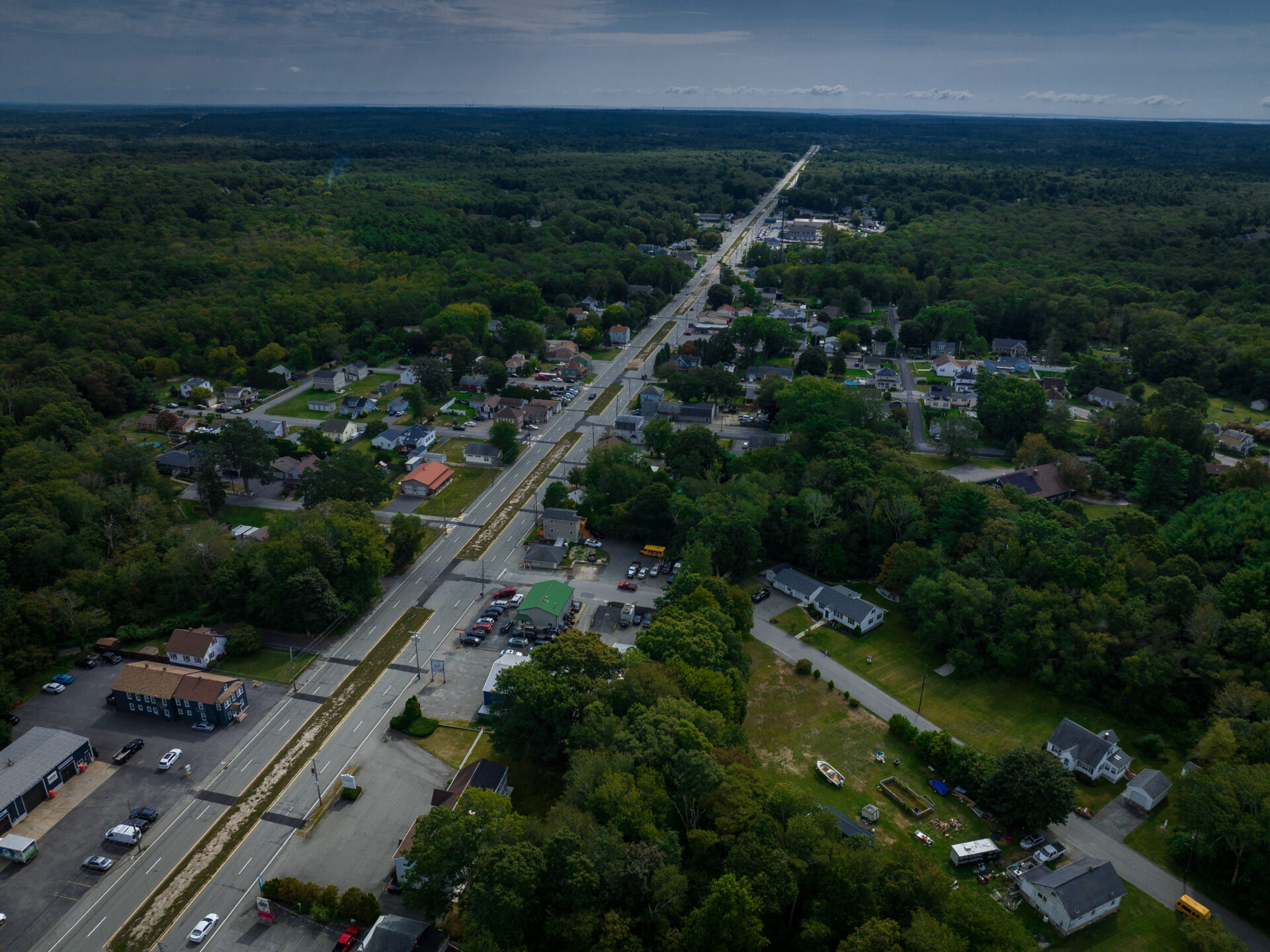 An aerial view of a divided four-lane highway lined with parking lots and strip malls cutting a straight line to the horizon through an otherwise wooded landscape.