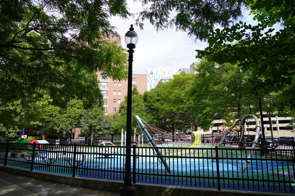 A view of a playground in a city park surrounded by mature trees. A handful of high-rise buildings are visible in the distance.