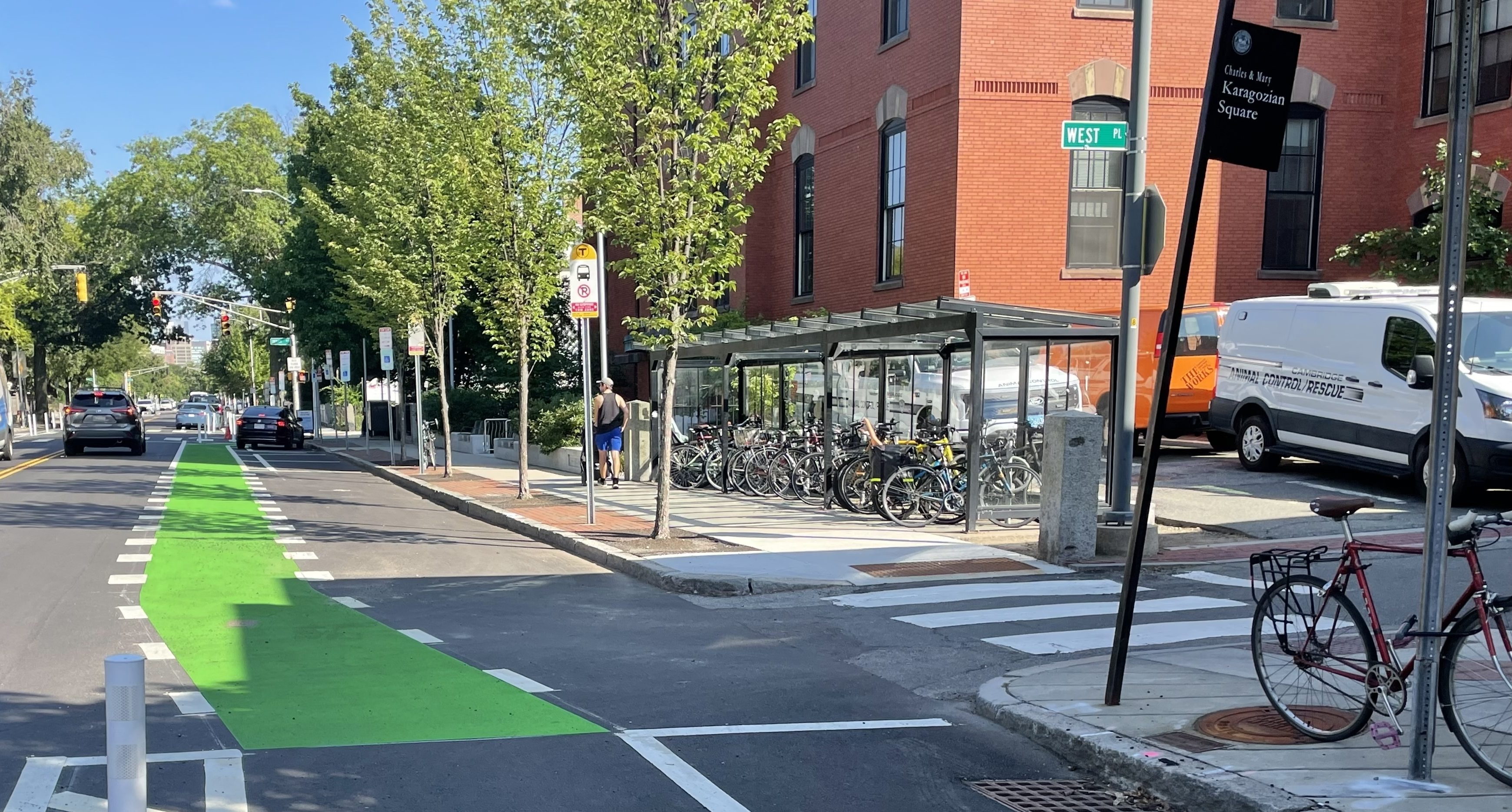 A glass bus shelter is full of parked bikes next to an older brick building (right) and an MBTA bus stop (left). In the street next to the bus stop is a bright green-painted bike lane.