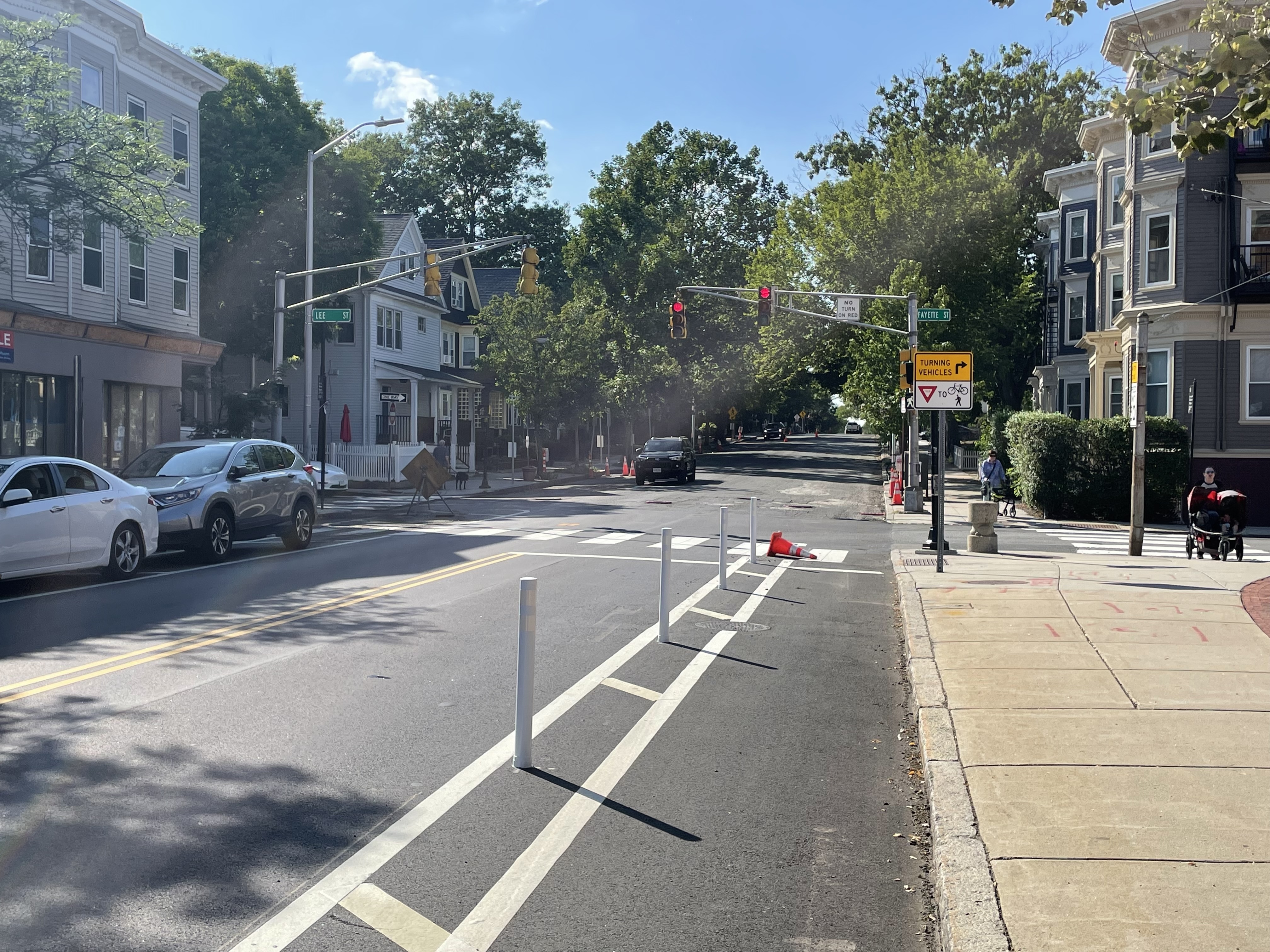 A protected bike lane runs next to white plastic bollards on a two-lane city street with parked cars on the opposite side. A traffic signal in the middle distance has a street sign that says "Lee St" on the left, and "Fayette St" on the right.