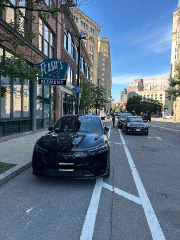 A low-resolution photo of a black sedan parked illegally in a bike lane next to an empty parking space in the legal parking lane. The street is lined with multi-story buildings and a hanging neon sign immediately to the left of the illegally parked car says "Flash's Element"