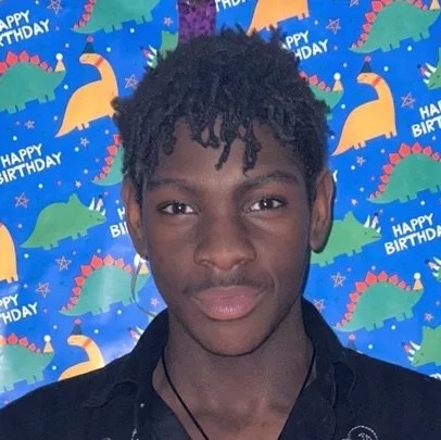 A young Black man with a slight smile stands in front of a "happy birthday" banner decorated with dinosaurs.