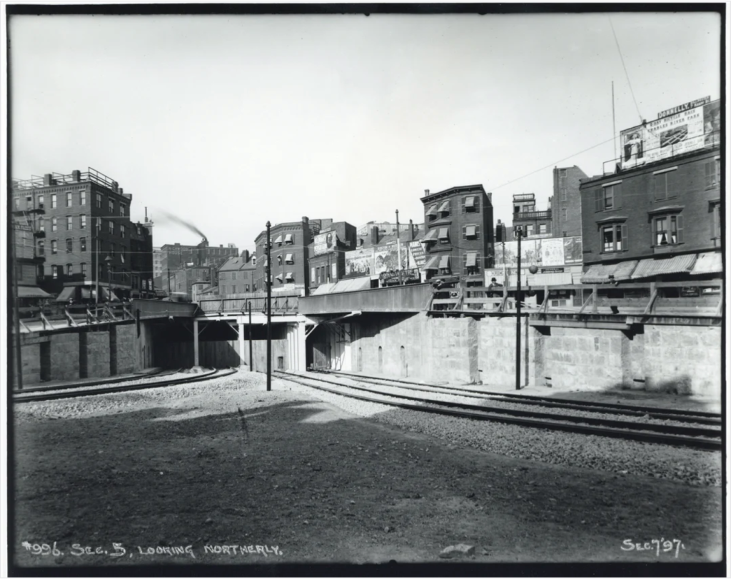 A black-and-white image of a subway portal with tracks leading into a tunnel. Old 3- and 4-story brick buildings line the streets above the subway portal.