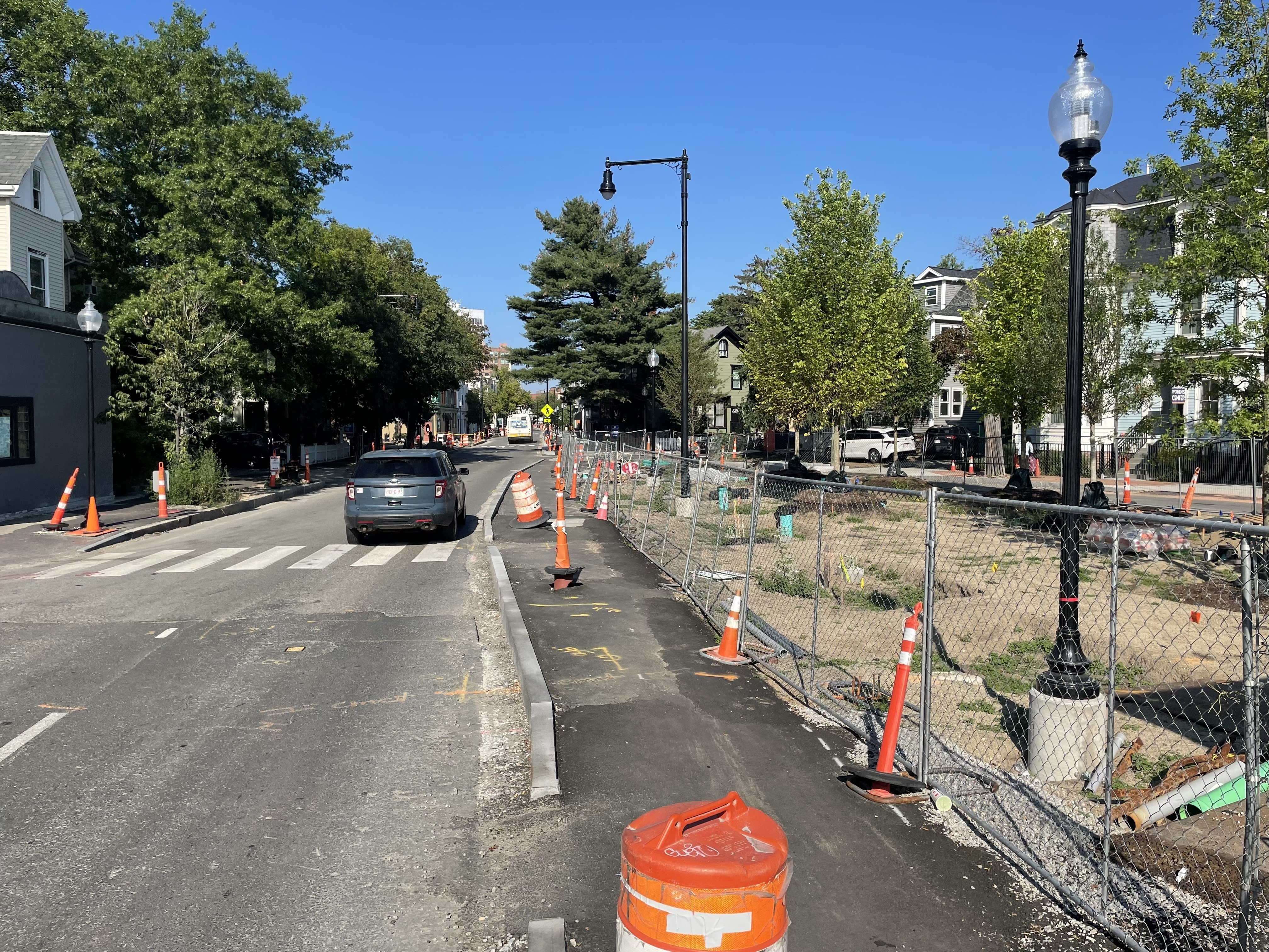 A street under heavy construction with an asphalt sidewalk/bikeway running through the center of the image and a park under construction with new trees in a dirt lot to the right. 