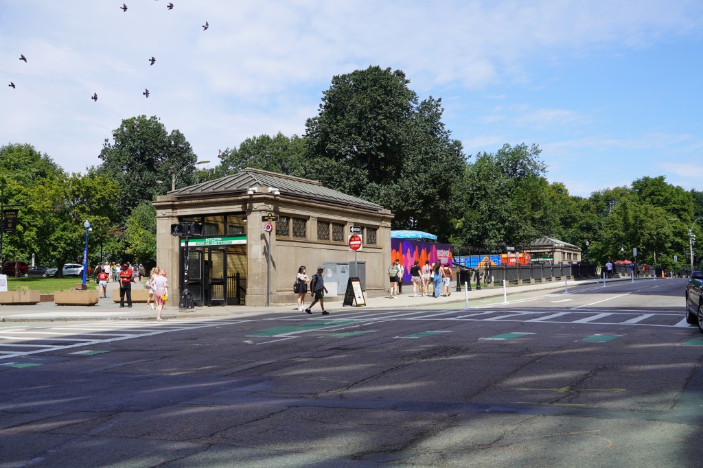 Two 19th-century masonry structures with pitched roofs in a park with mature leafy trees. Signs on the structures read "BOYLSTON" on a green background. Groups of pedestrians surround the structures on the sidewalk of a city street in the foreground.