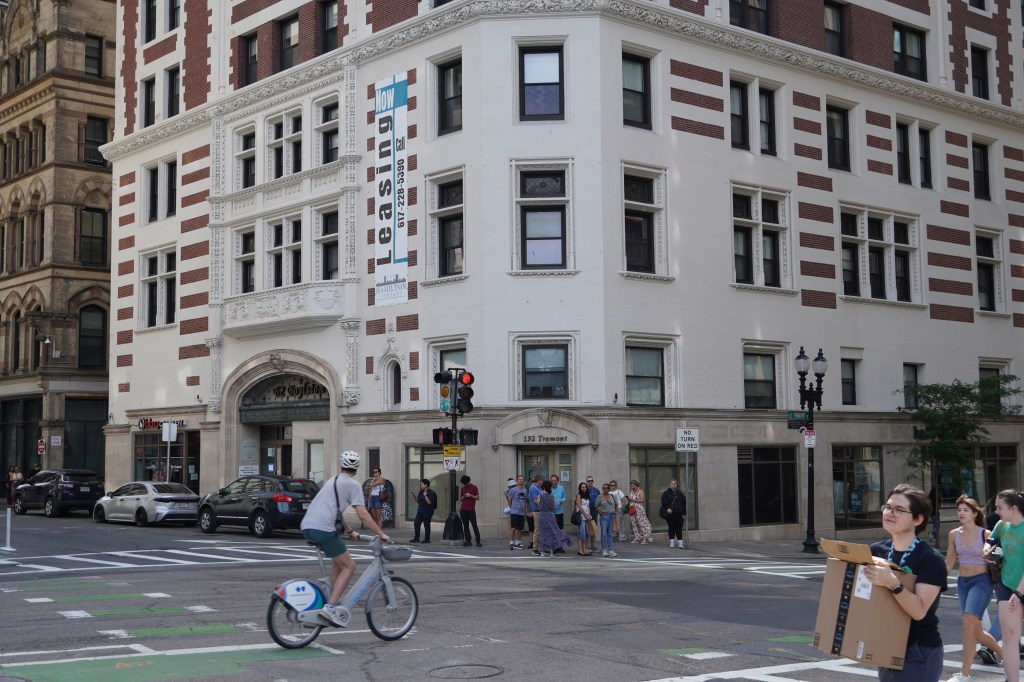 A view of a city intersection with a person riding a bike and crowds of pedestrians in the foreground. In the background is a large 19th-century masonry building with a tall "Leasing" banner on its side.