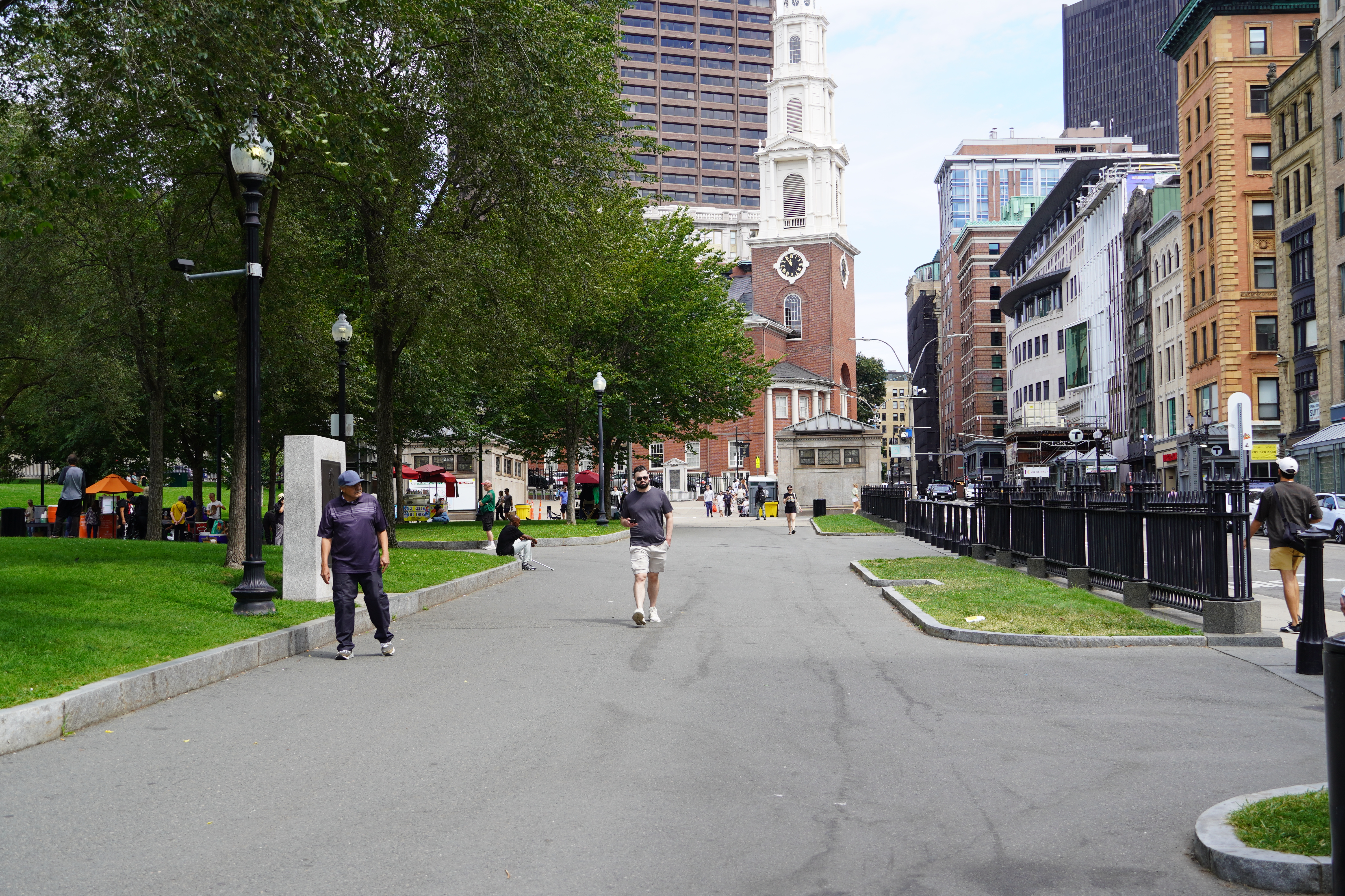 A wide walkway in a leafy park in downtown Boston. At the end of the walkway is the steeple of a large church with a taller skyscraper beyond; on the right is a street lined with mid-rise buildings in a mix of architectural styles.