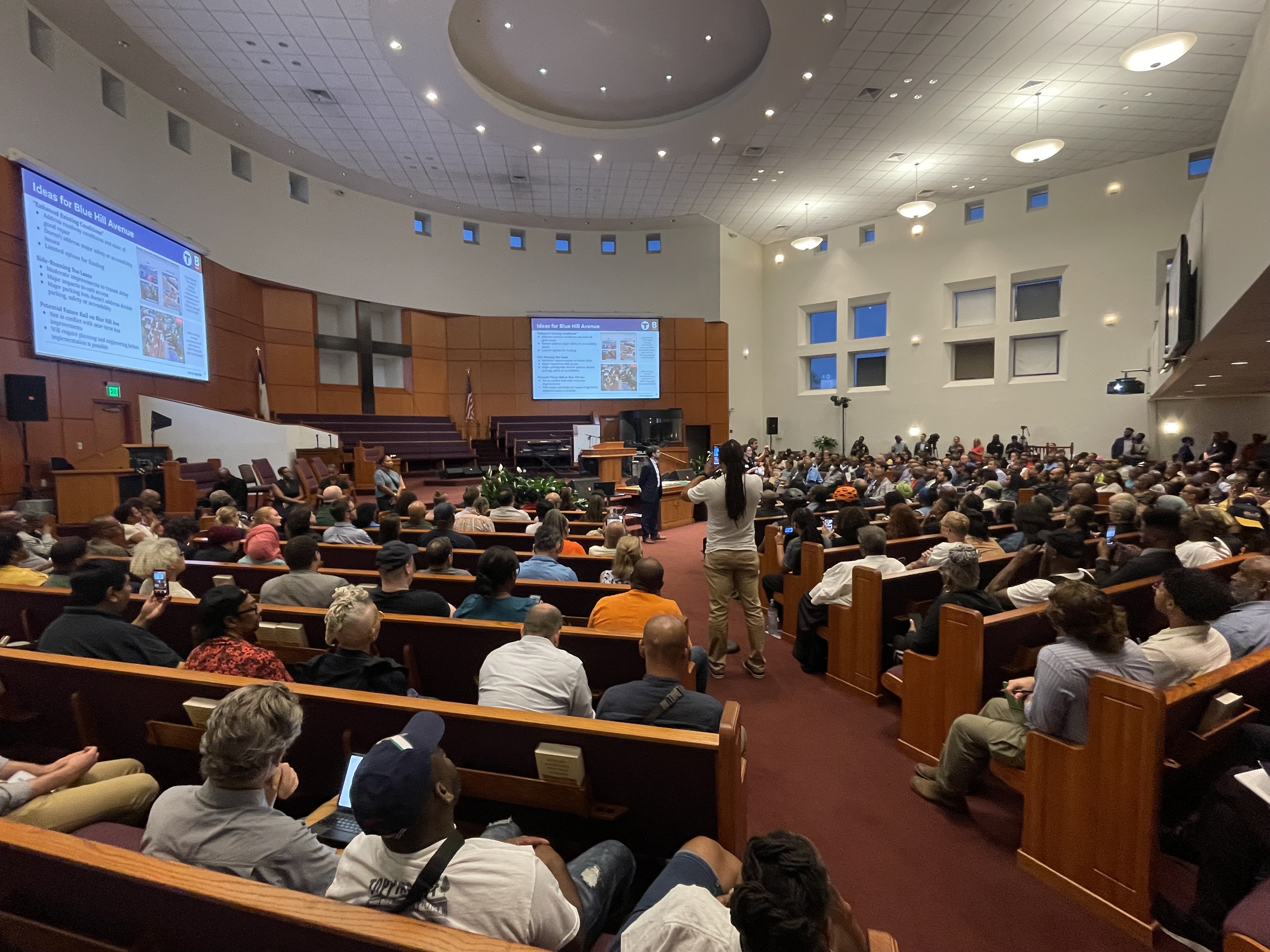 The interior of a modern circular church sanctuary with pews full of people facing two speakers in the center of the room. Behind them on a large screen on the wall is a presentation slide titled "Ideas for Blue Hill Avenue"