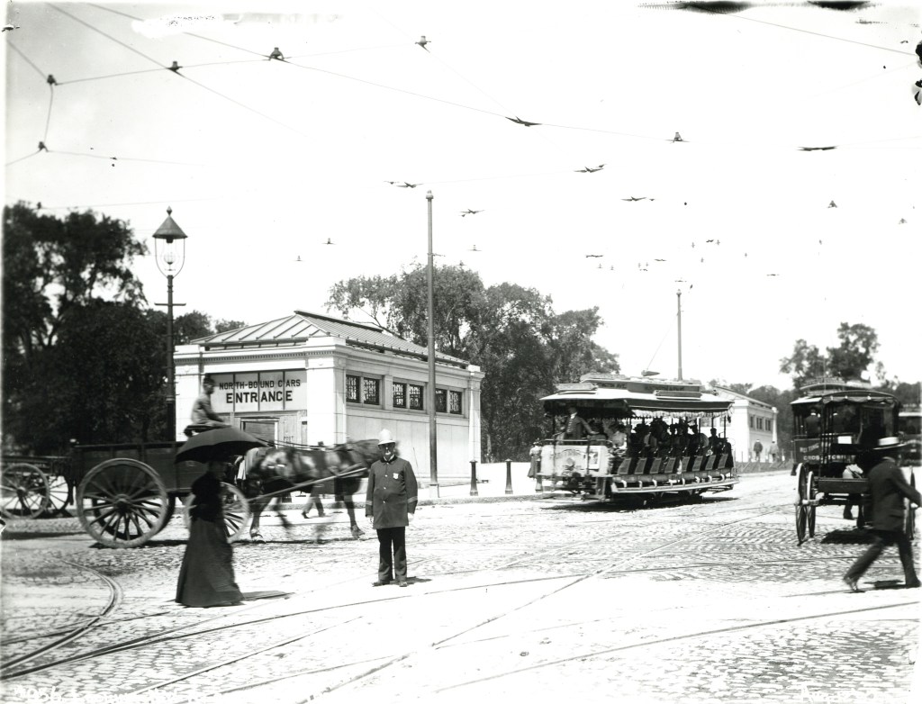 A black and white photo of a 19th-century city street with several crowded open-sided streetcars, a horse-drawn cart, and a police office directing traffic. In the background are two masonry structures with pitched roofs in a park with mature leafy trees. A sign on the closer structure reads "NORTH-BOUND CARS ENTRANCE"
