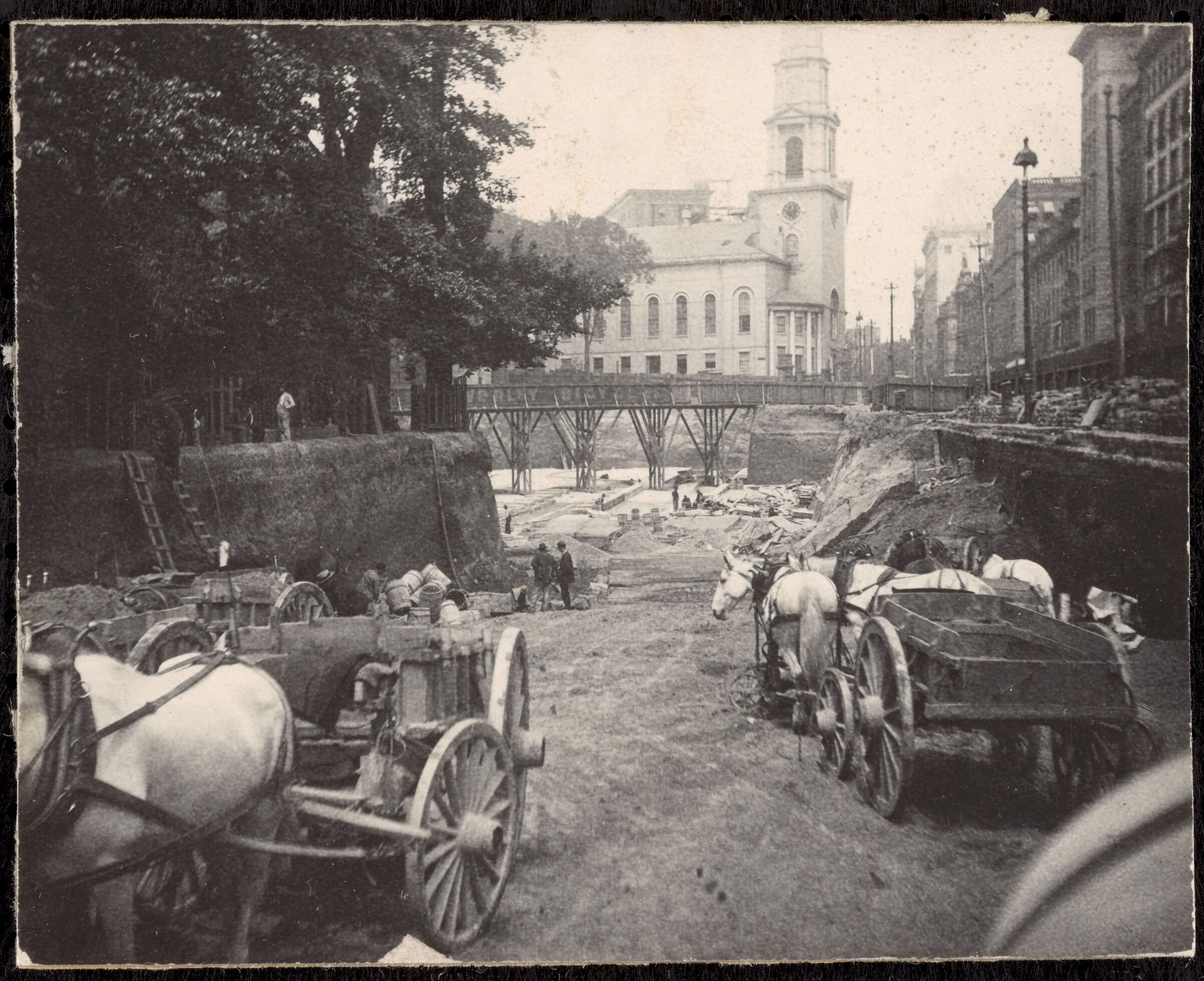 A black-and-white image of a major excavation pit in a 19th-century city. In the background is a tall church, and on the right edge are a row of tall multi-story masonry buildings. In the foreground are several horse-drawn carts descending an earth ramp into a massive, deep hole. On the left is a park with mature trees. In the middle distance, in the hole but in front of the church, is a tall wooden bridge spanning the pit.