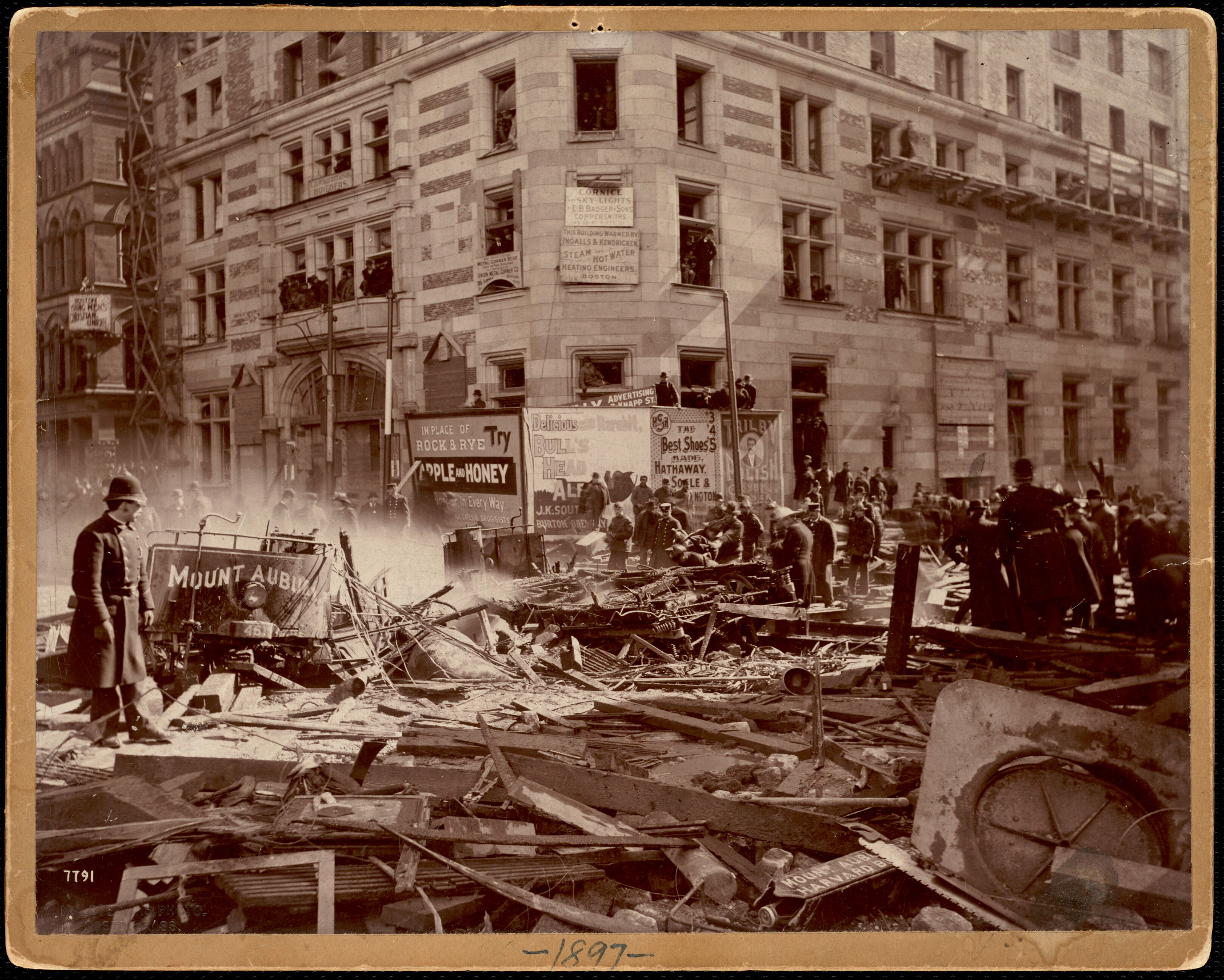 A sepia-toned black-and-white photo of a city street covered in broken timbers and other debris in the aftermath of a major explosion. In the background is a large multi-story masonry building with many broken windows.