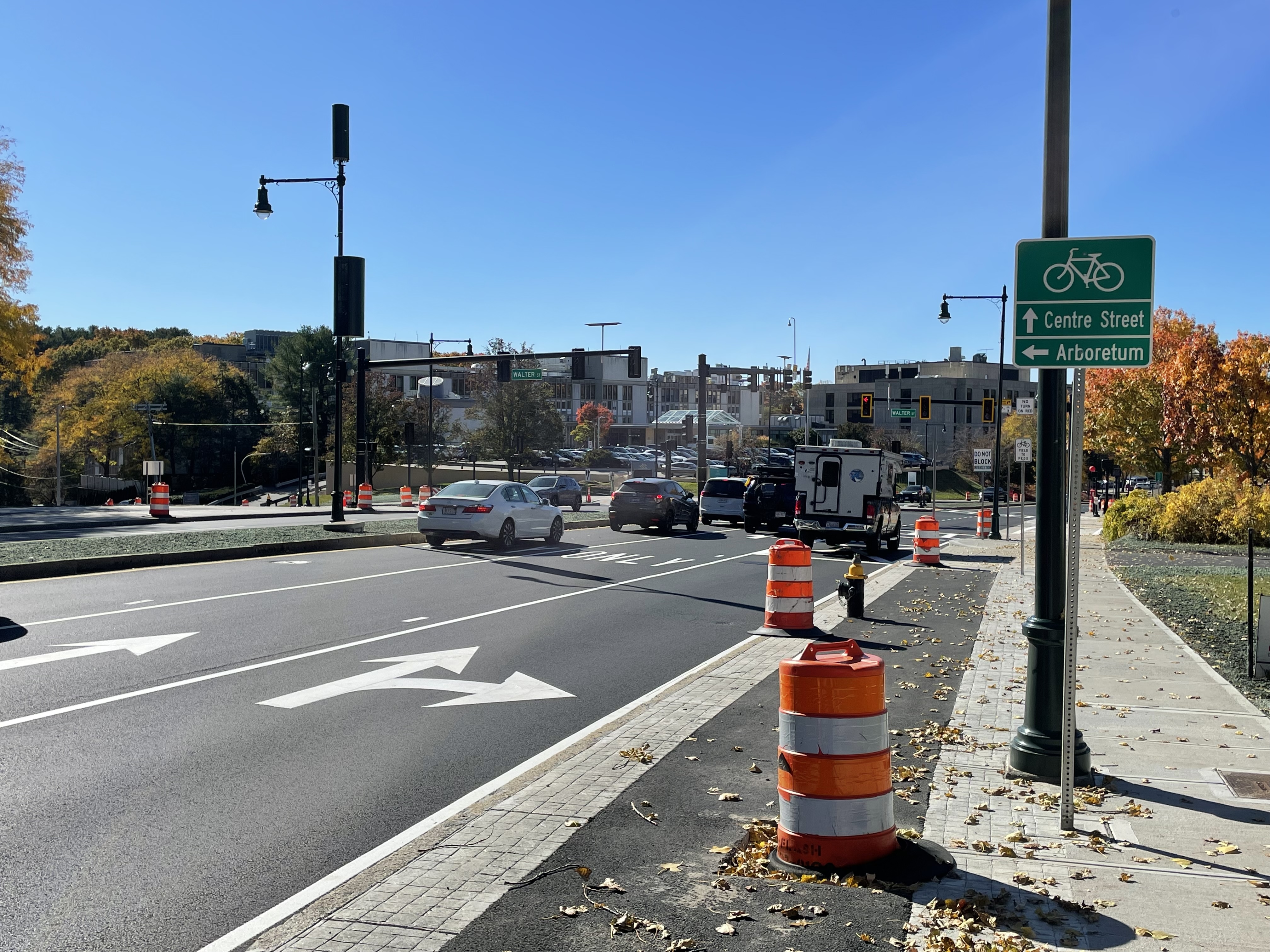 A view of a new sidewalk with a sidewalk-level protected bike lane lined with orange construction barrels. A green bike route sign in the foreground at the edge of the sidewalk reads "Centre Street (straight arrow), Arboretum (left arrow)". In the distance is a traffic light with five waiting cars and trucks.