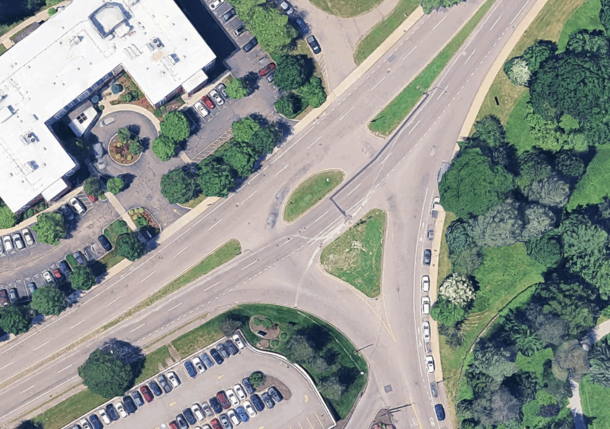 An animated GIF showing a satellite view of a wide Y-shaped intersection next to a park at right, a large building in upper left, and a parking lot in lower left. The larger street runs diagonally from lower left to the upper edge, with green islands in the middle of the roadway. The smaller roadway comes in from the bottom edge and intersects the large road at a green splitter triangle. The image includes an overlay of black lines labelled "new curbline" which outlines a new, narrower intersection with a basic T-junction replacing the green triangle in the center of the "y". Subsequent frames of the animation show new curblines being built, and the plans for new sidewalks, bike lanes, and crosswalks in a new T-shaped intersection that occupies a much smaller footprint.