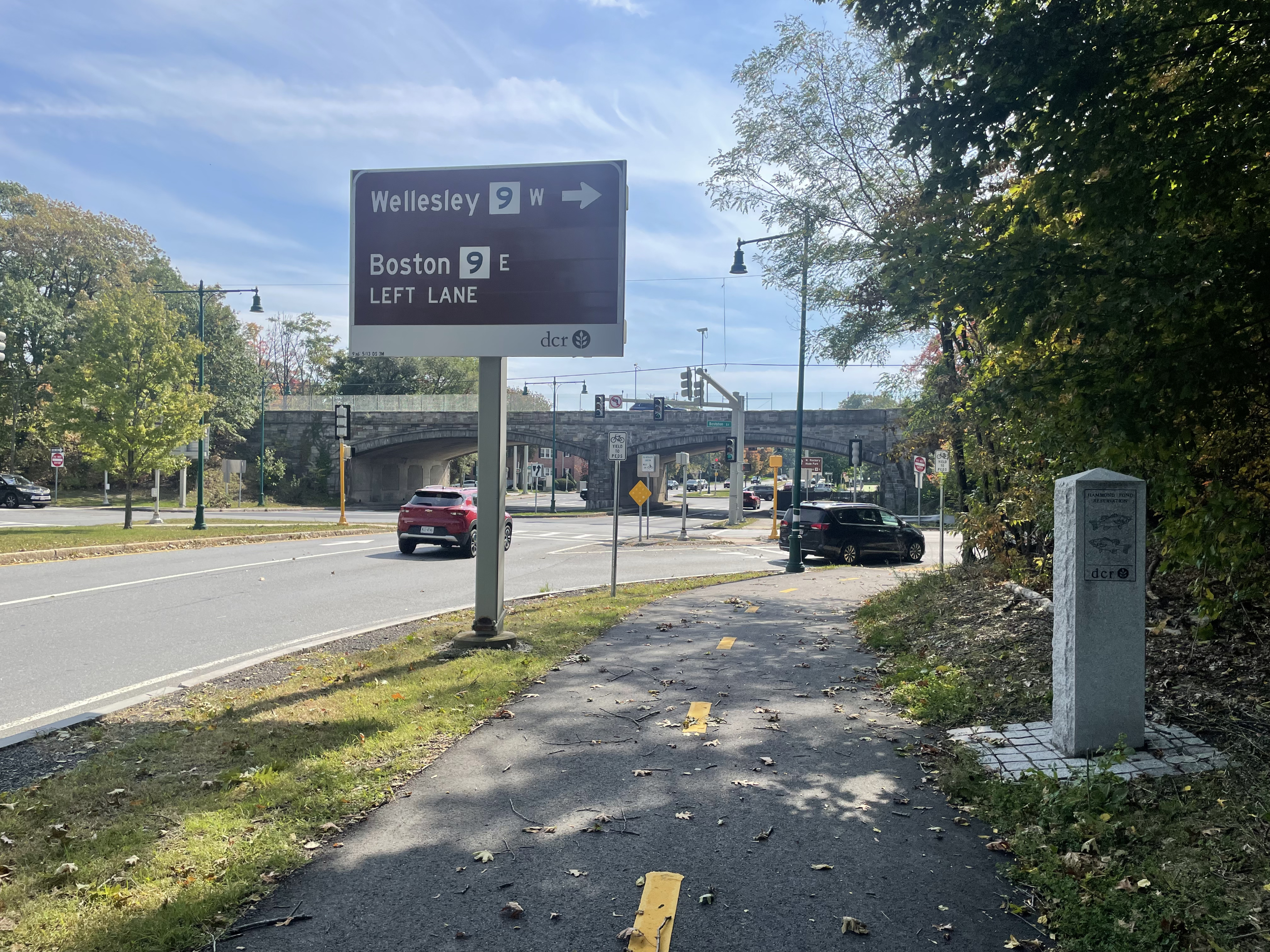 A bike path runs between a granite signpost and a large brown highway sign that reads "Wellesley Route 9 W (right arrow)" and "Boston 9 E left lane". Beyond the sign is a large intersection under a stone arch highway viaduct.