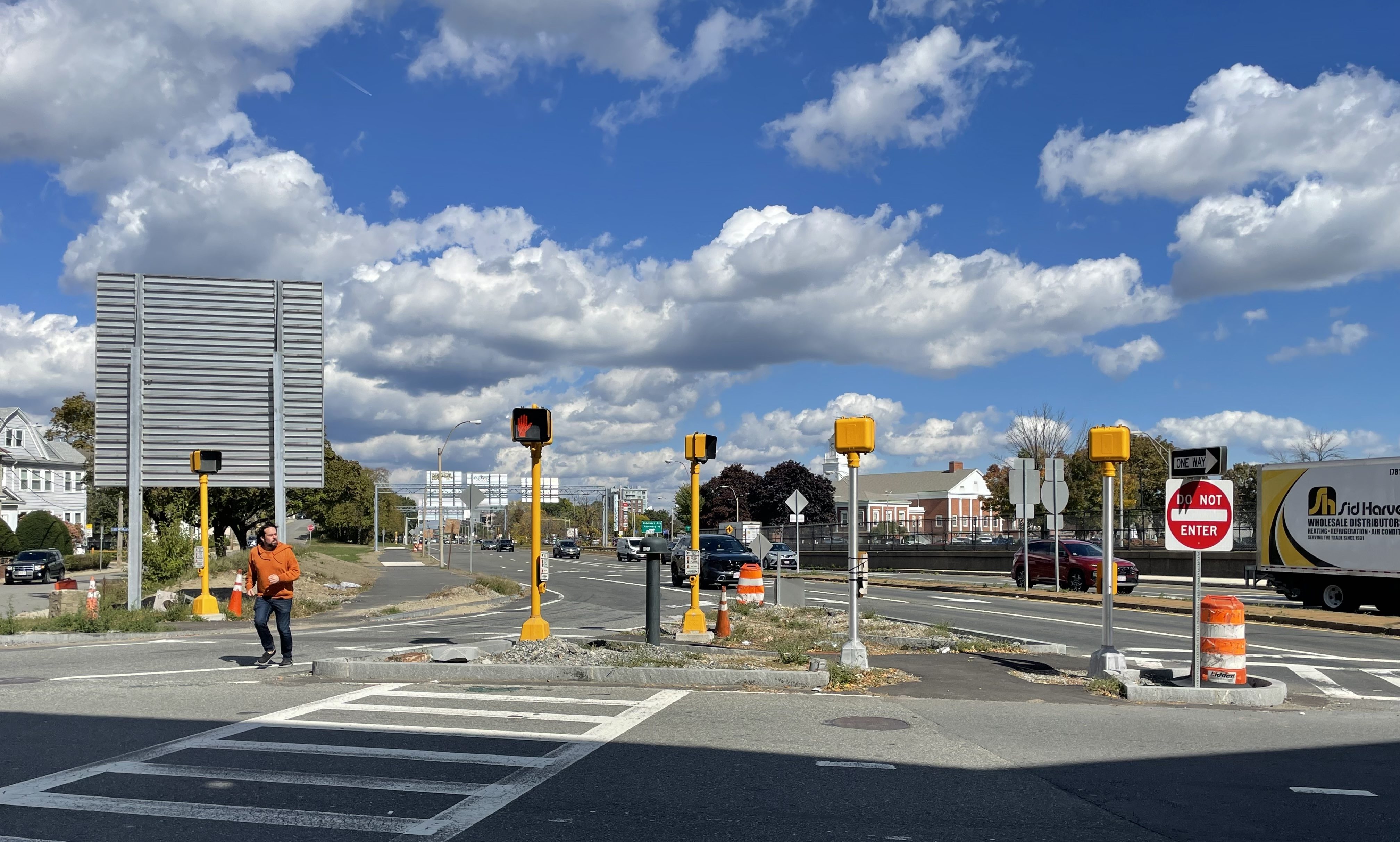 A triangular traffic island is studded with four pedestrian signals in the middle of a wide intersection between two wide multi-lane streets. To the left, a man in a red jacket is in the middle of jogging across the multi-lane roadway in the foreground. Behind him is the grey back side of a large highway sign, approximately 12 feet wide and tall.