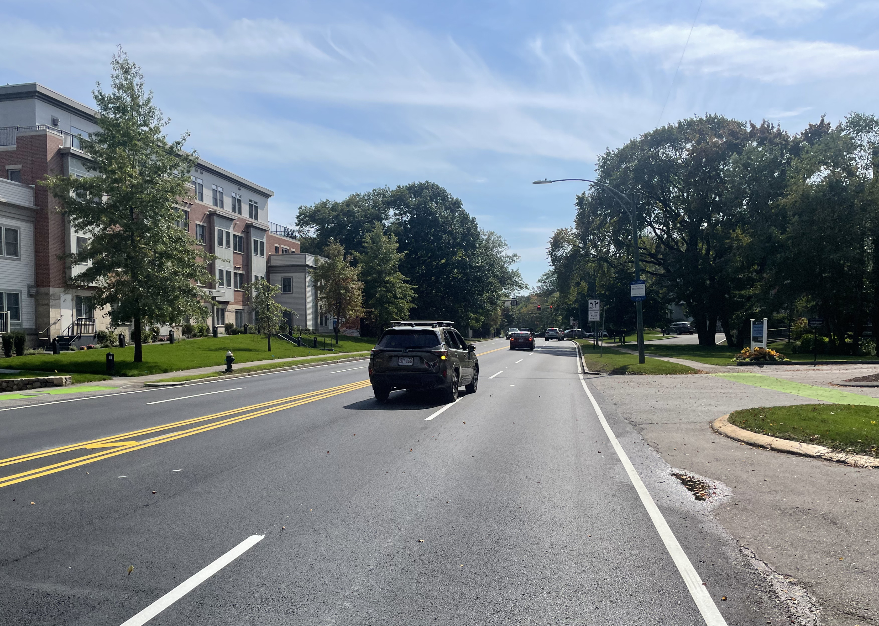 A recently paved wide 4-lane roadway lined with mature trees and (at left) a 4-story apartment building.