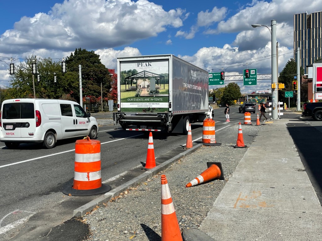A van and a large box truck stop at a red light on a wide multi-lane street lined with orange construction cones while two people walk across a crosswalk in the middle distance under the traffic light. In the foreground a strip of gravel lies parallel to a concrete sidewalk. Beyond the traffic light are two large green highway signs. One reads "28 NORTH Malden" above two downward pointing arrows, and the other reads "38 Medford Boston" above one downward pointing arrow.