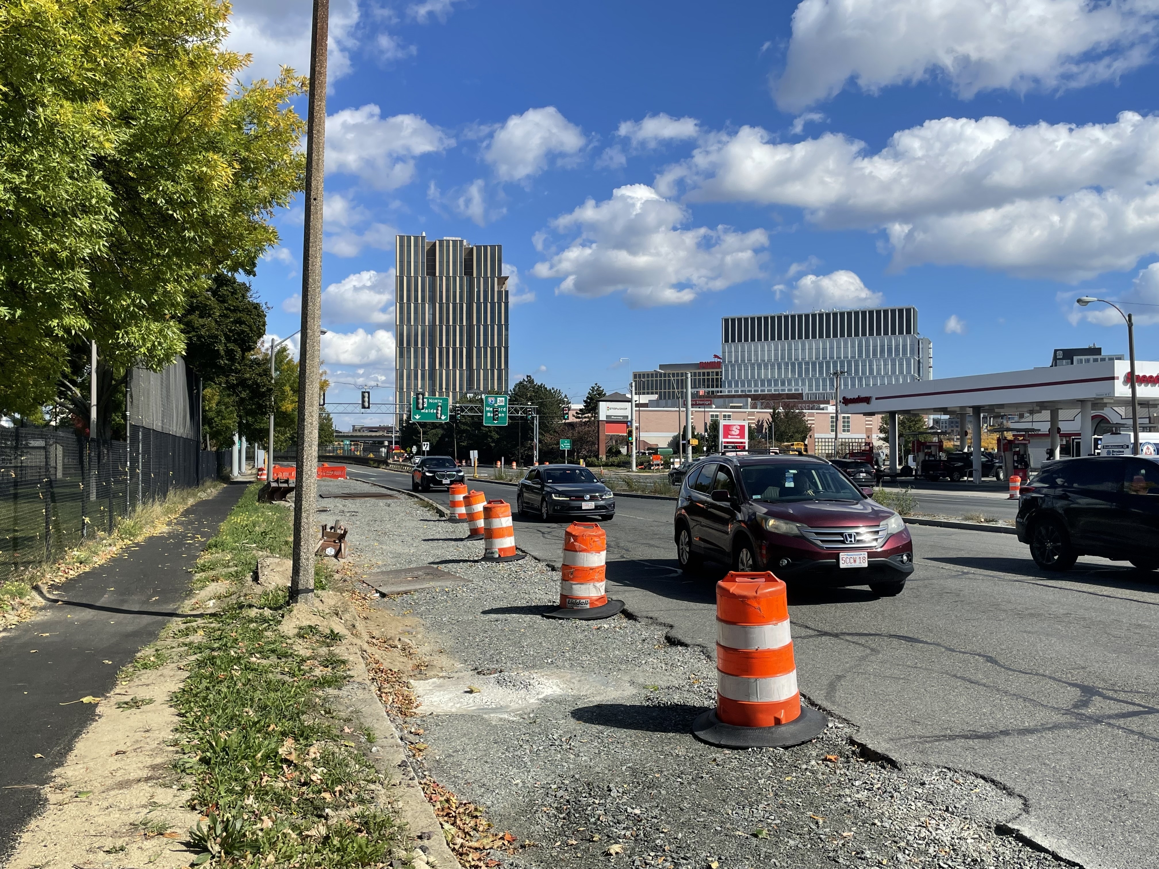 A photo of a wide multi-lane street lined with orange construction cones (right) next to a park with trees (at left). In the foreground a strip of gravel lies parallel to an asphalt sidewalk. 