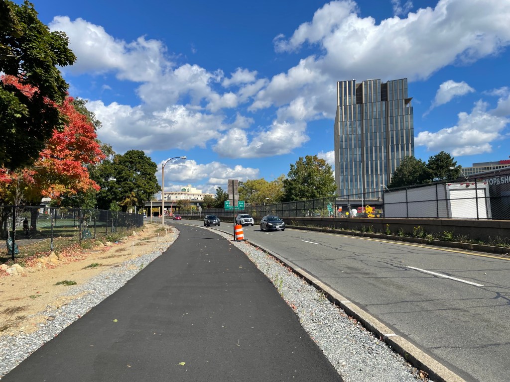 A newly-paved 10-foot pathway runs next to a city park (left) and a two-lane roadway with three cars. In the distance is a high-rise building.