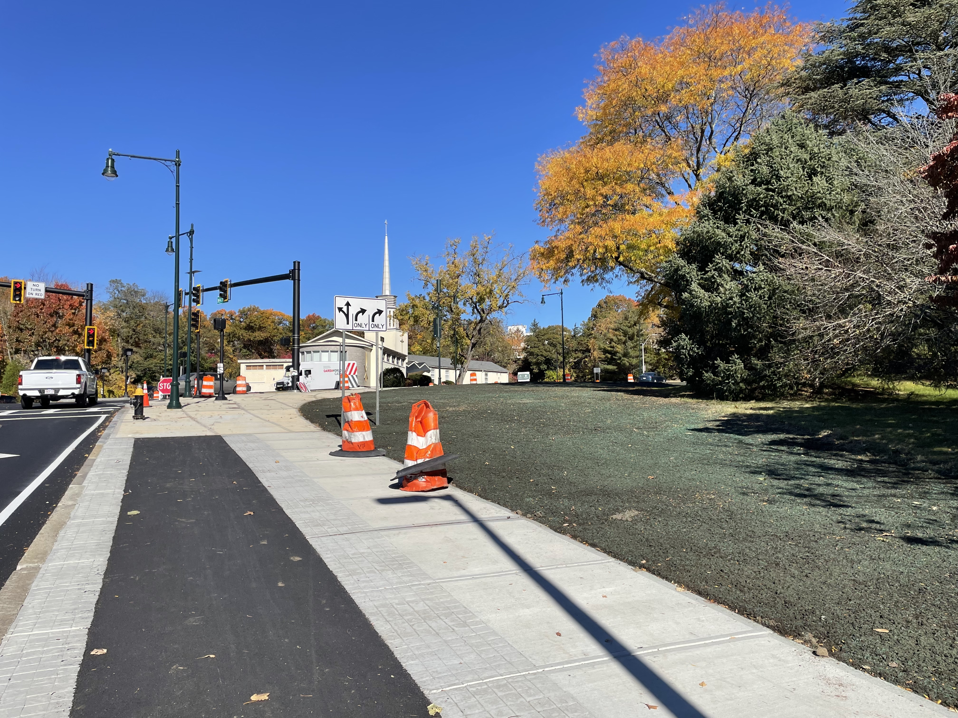 A newly-seeded crescent of lawn alongside a recently-constructed sidewalk with a handful of orange construction barrels. To the right are several mature trees with yellow leaves. On the left is a traffic light.