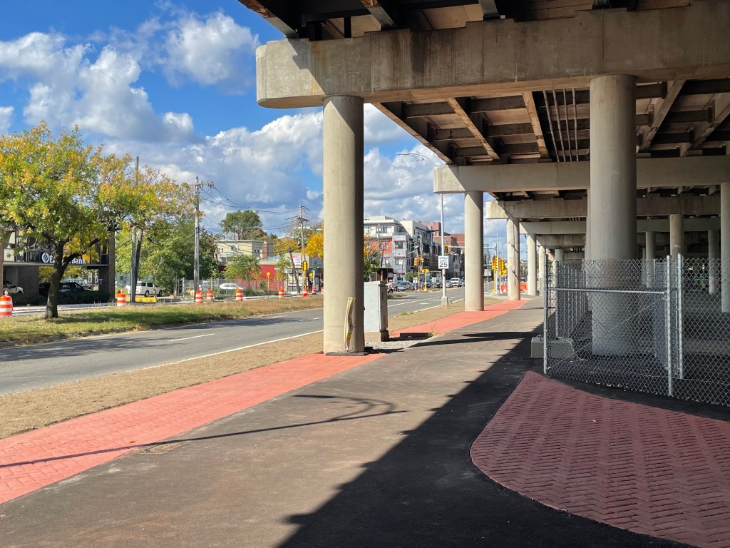 A wide asphalt path runs under the edge of an elevated highway viaduct next to a wide multi-lane surface road with city buildings on the other side. A sign in the middle distance reads "SPEED LIMIT 25".