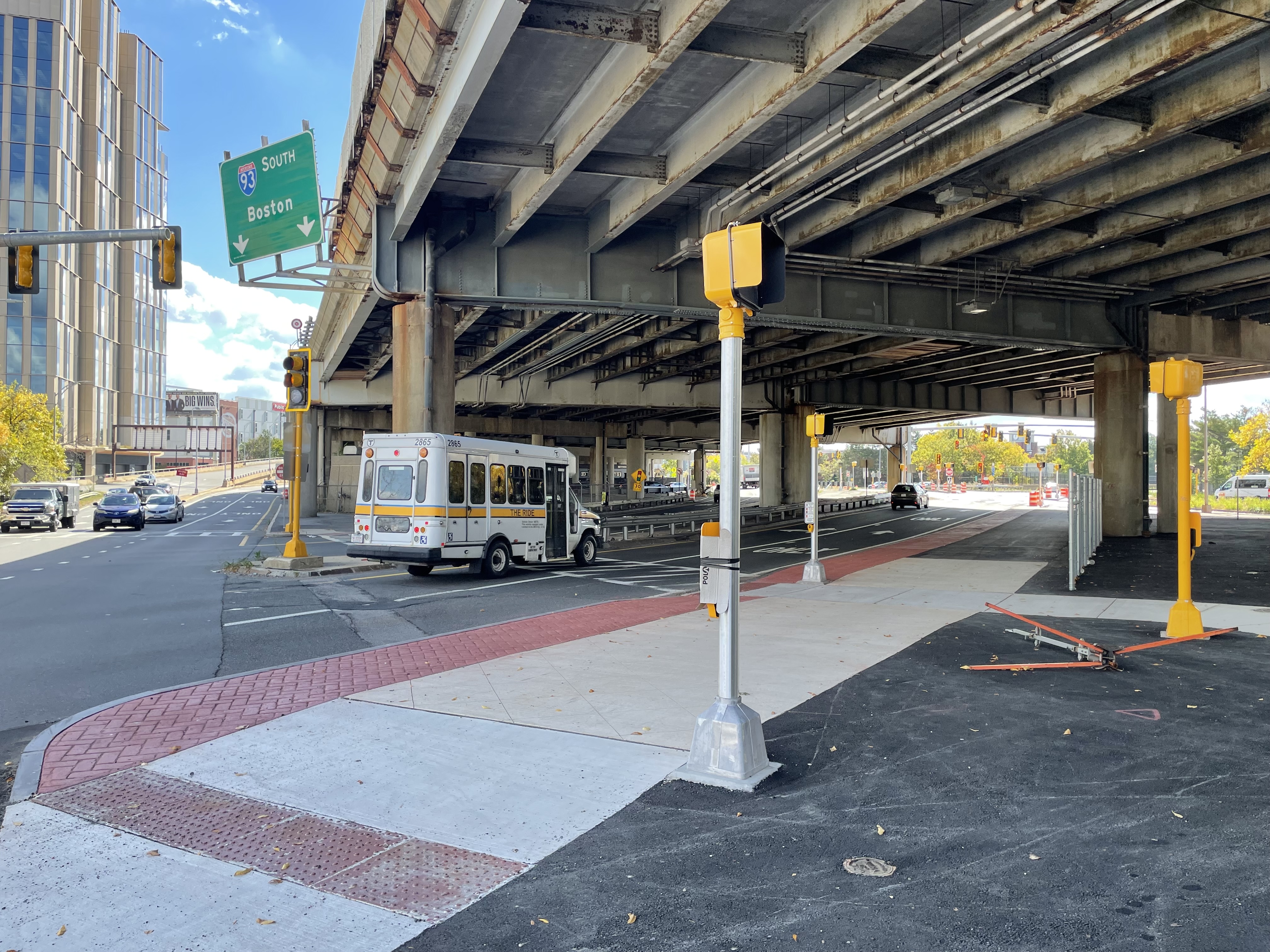 A wide, 15-foot-side concrete sidewalk runs diagonally under a wide, aging highway viaduct next to a multi-lane roadway where an MBTA The Ride van is driving A green highway sign above reads "93 South Boston" above two downward-pointing arrows. On the left edge of the photo is a high-rise building.
