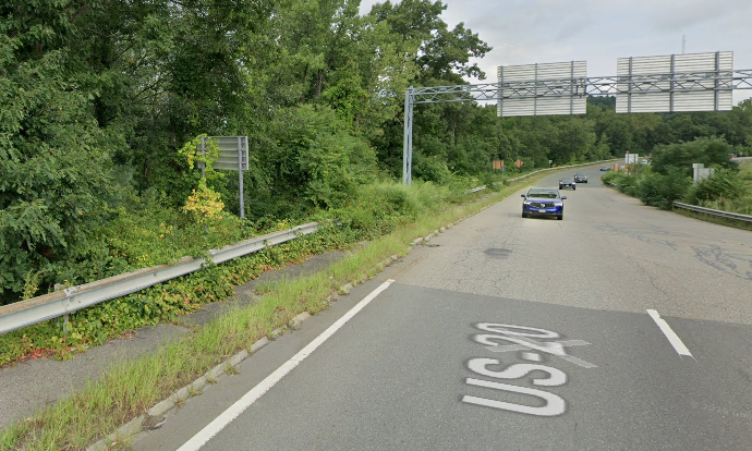 A Google Street view image of a wide highway ramp alongside a narrow, poorly-maintained and overgrown sidewalk next to a guardrail. 